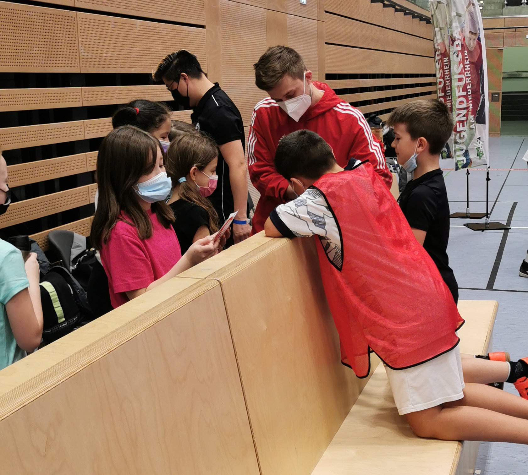 [{"clang_id":1,"value":"Gruppe Kinder mit Masken beugt sich über eine niedrige Holzbank in einer Turnhalle. Einige tragen Sportwesten. Im Hintergrund eine Wand mit Schriftzug."}]