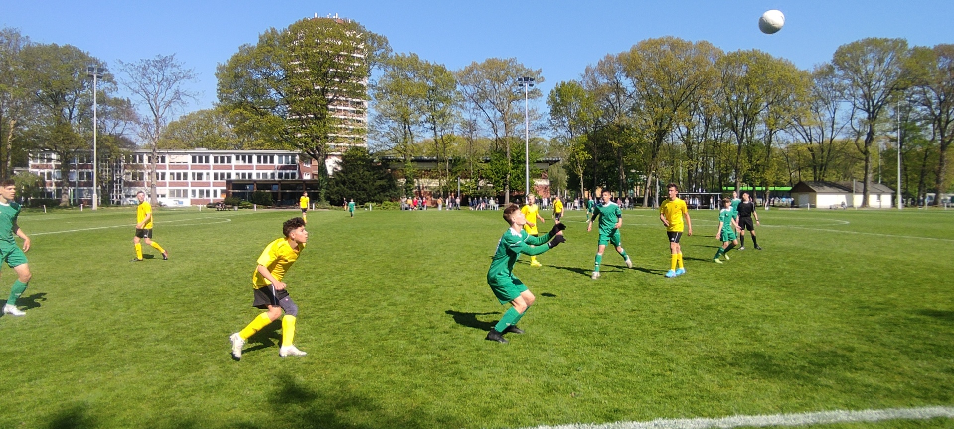 [{"clang_id":1,"value":"Fußballspiel auf grünem Rasenfeld. Spieler in grünen und gelben Trikots, einige in Bewegung, ein Ball in der Luft. Im Hintergrund Gebäude und Bäume."}]