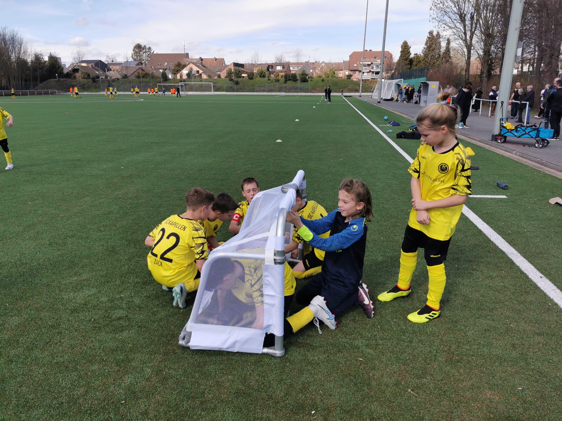 [{"clang_id":1,"value":"Fußballfeld. Eine Gruppe Kinder in gelben Trikots umringt ein kleines Fußballtor, einige knien davor. Im Hintergrund weitere Spieler und Zuschauer."}]