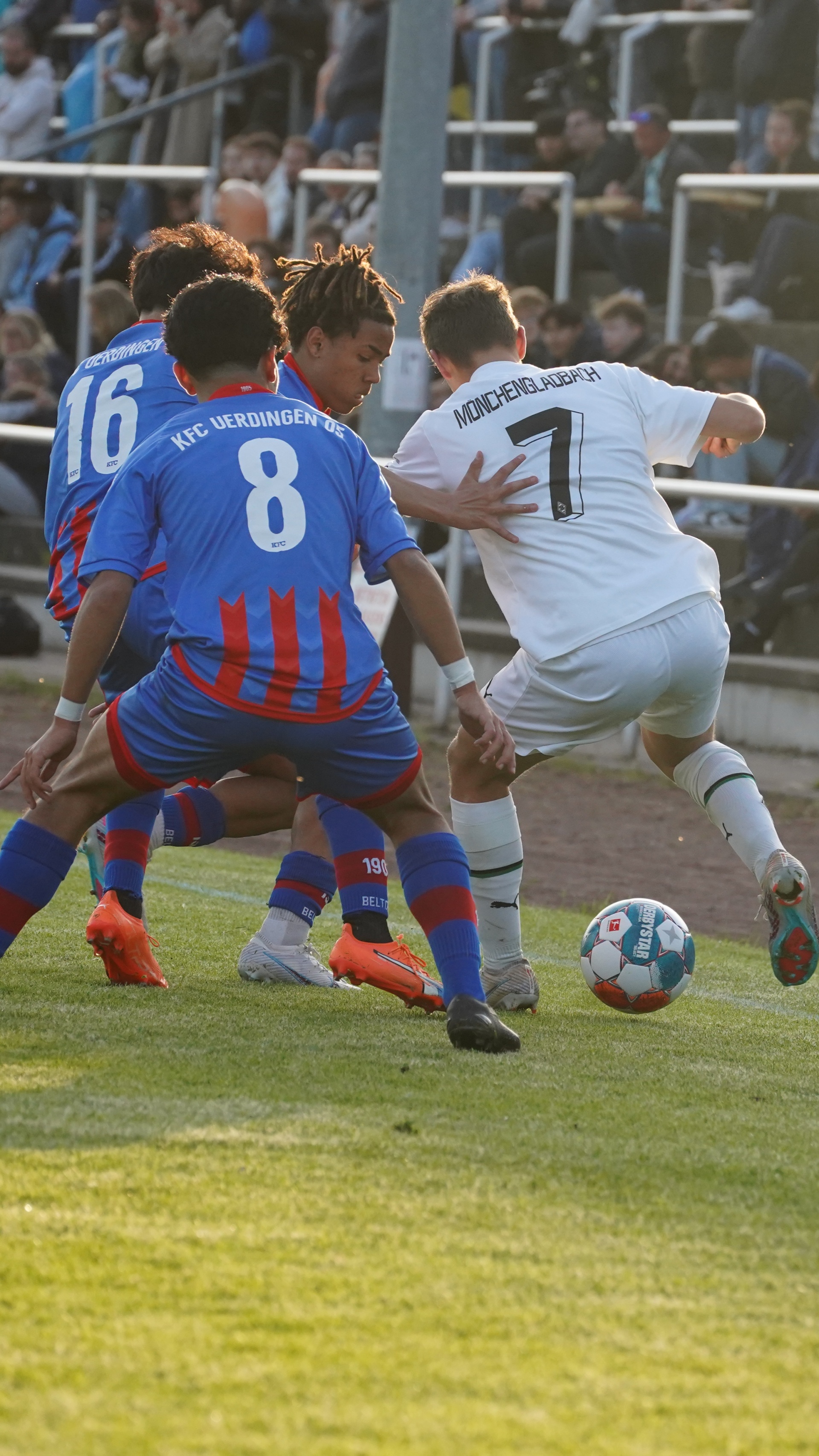 [{"clang_id":1,"value":"Fußballspiel: Drei Spieler kämpfen um den Ball. Zwei tragen blaue Trikots mit den Nummern 8 und 16, einer ein weißes Trikot mit der Nummer 7. Im Hintergrund Zuschauer."}]