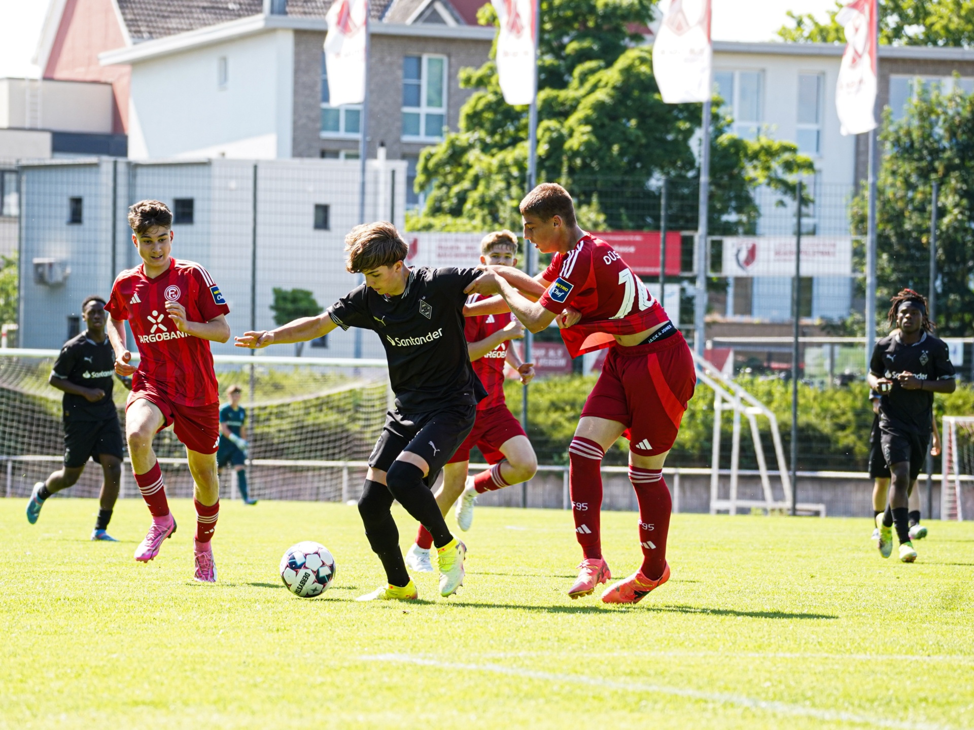 [{"clang_id":1,"value":"Fußballspiel: Drei Spieler kämpfen um den Ball auf grünem Rasen. Links und rechts rot gekleidete Spieler, in der Mitte ein Spieler mit schwarzem Trikot. Im Hintergrund Gebäude und Bäume."}]