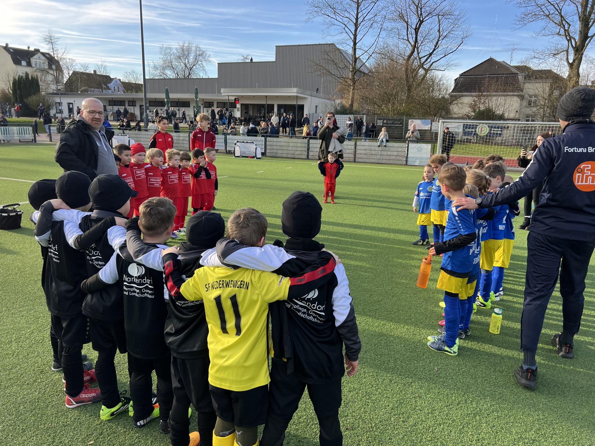 [{"clang_id":1,"value":"Fußballteam im Huddle auf grünem Rasen. Spieler tragen blaue und schwarze Trainingsanzüge, einige gelbe Westen. Spiegelung im Wasser erkennbar."}]