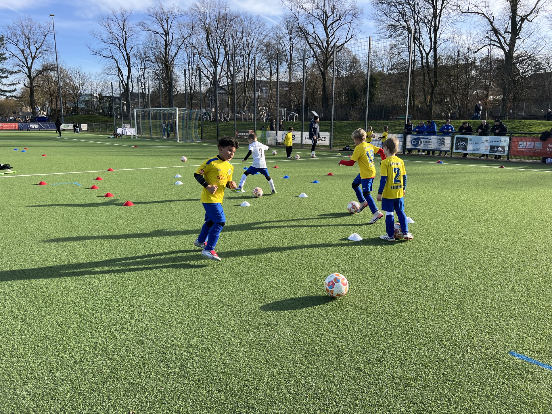 [{"clang_id":1,"value":"Fußballtraining: Kinder in gelben und dunklen Trikots üben Dribbling um Hütchen auf einem grünen Kunstrasenplatz. Im Hintergrund Bäume und Zuschauertribüne."}]