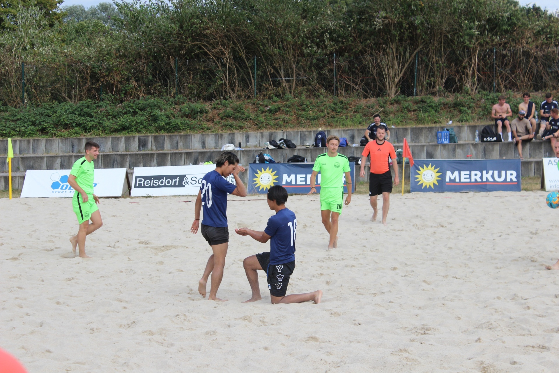 [{"clang_id":1,"value":"Strandfußballspiel: Vier Spieler in farbigen Trikots auf sandigem Spielfeld, ein Spieler kniet. Im Hintergrund eine Zuschauertribüne mit Personen und Büschen."}]
