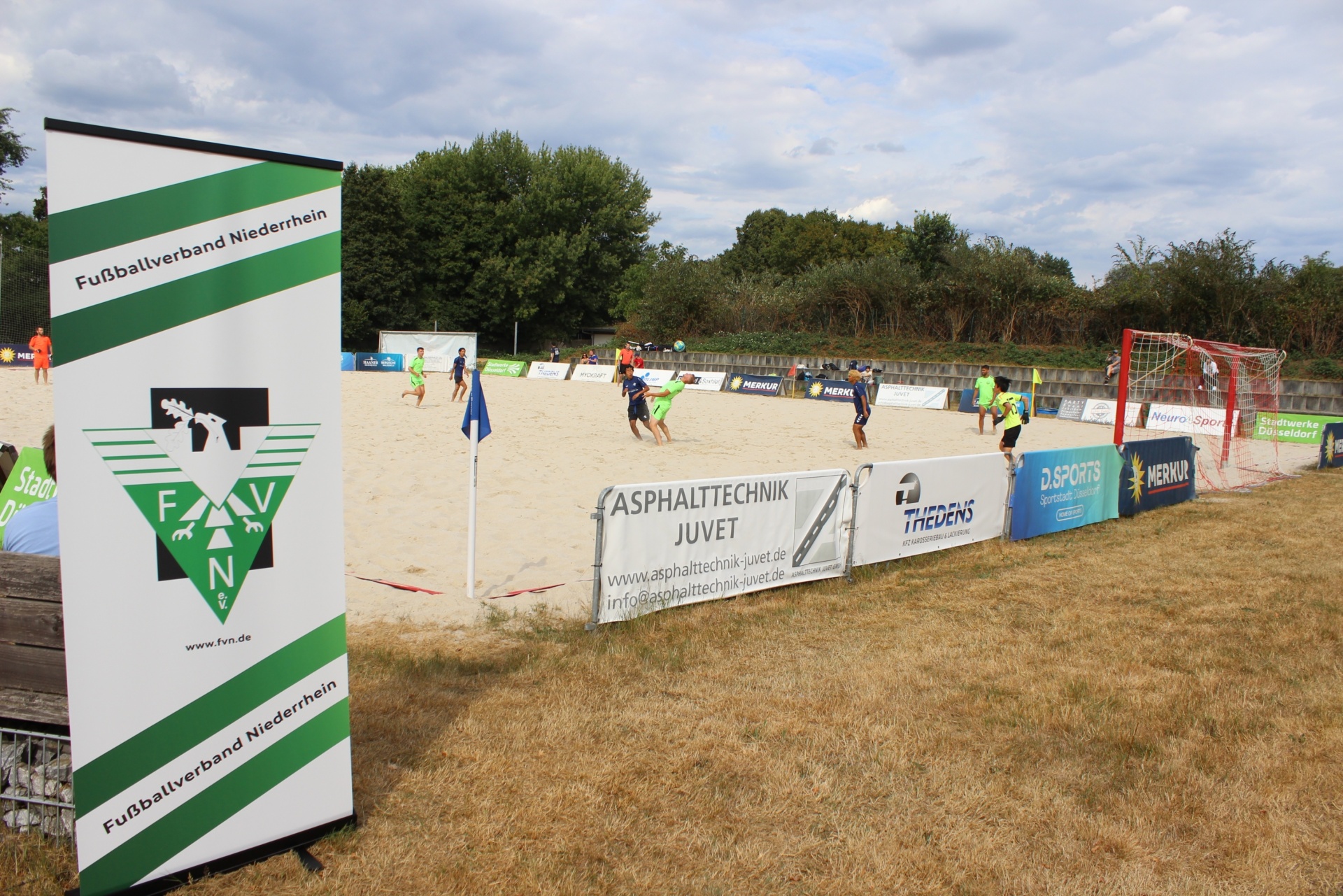 [{"clang_id":1,"value":"Beachvolleyballfeld mit Spielern und Werbebanden. Links Banner \"Fußballverband Niederhein\". Hintergrund: Bäume, Himmel."}]