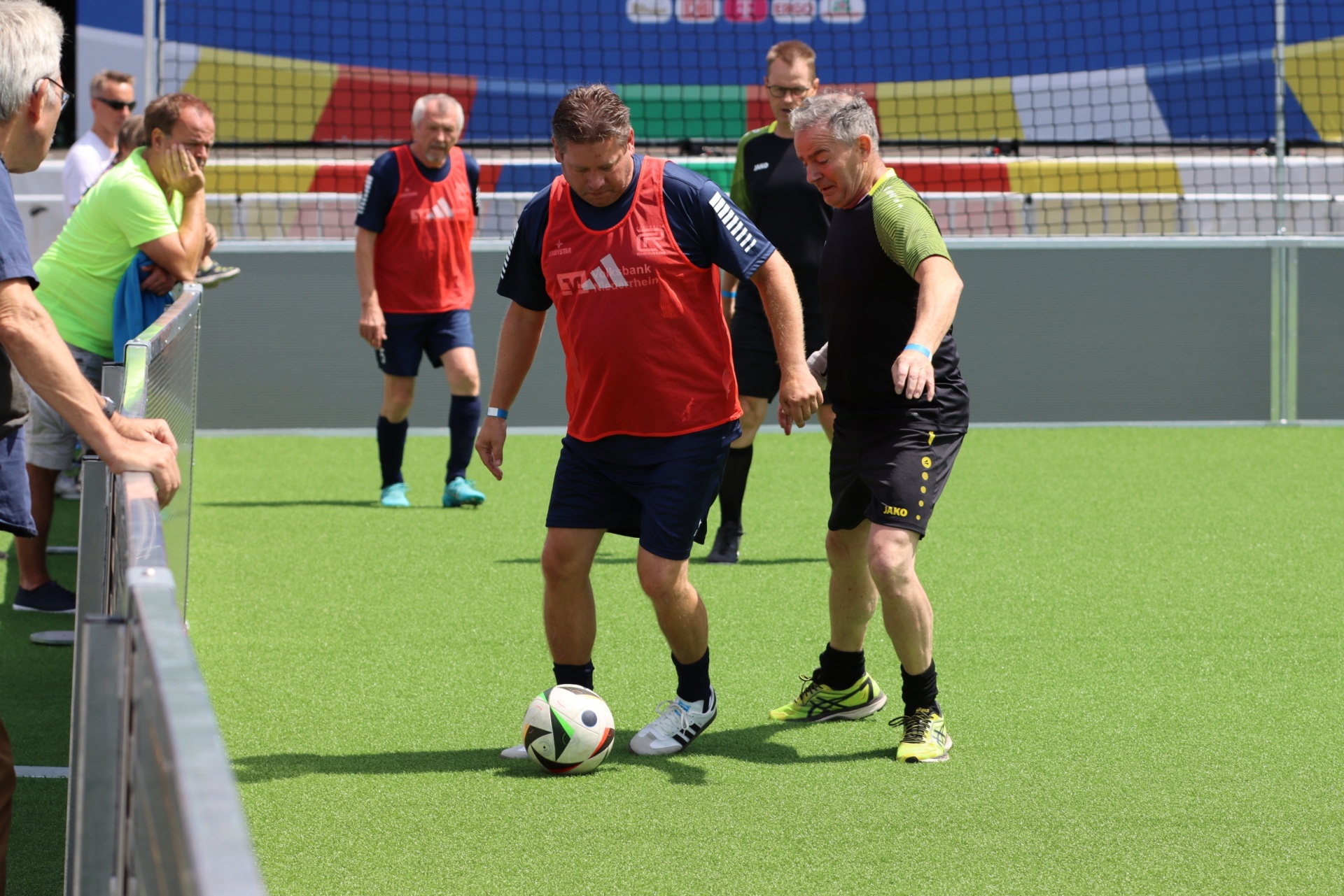 [{"clang_id":1,"value":"Zwei Männer spielen Fußball auf einem grünen Kunstrasenplatz. Im Hintergrund stehen weitere Personen und eine farbige Bande."}]
