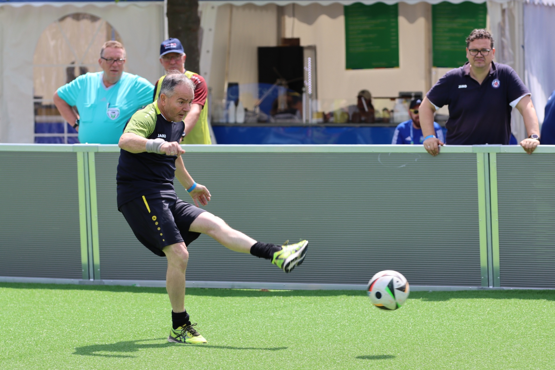 [{"clang_id":1,"value":"Mann tritt Fußball auf grünem Kunstrasen. Im Hintergrund steht eine Absperrung und Zuschauer unter einem weißen Zelt."}]