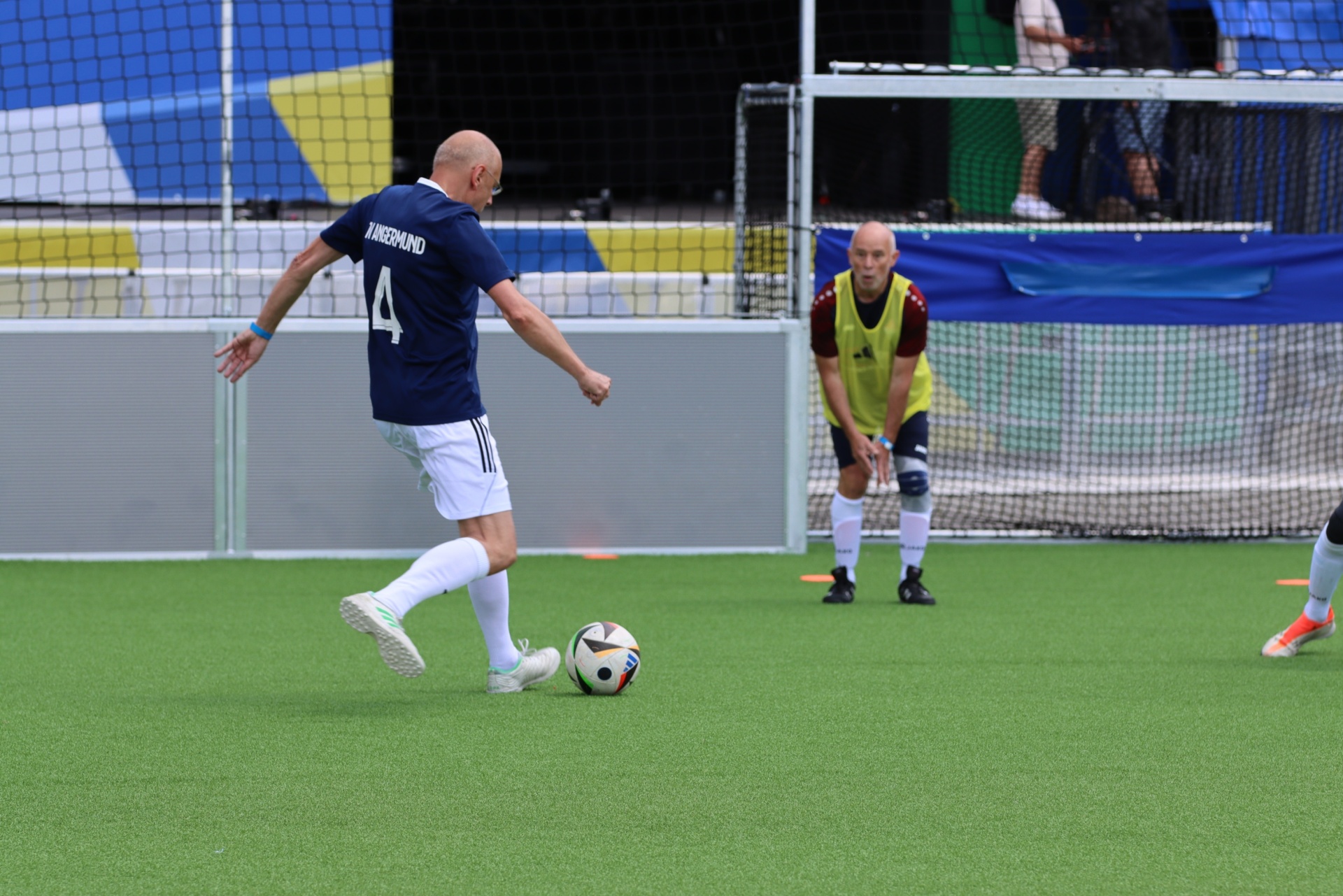 [{"clang_id":1,"value":"Fußballspieler mit Nummer 4 schießt Ball auf Tor, dahinter Gegenspieler in gelber Weste, grüne Spielfläche, Hintergrund mit Zuschauertribüne."}]