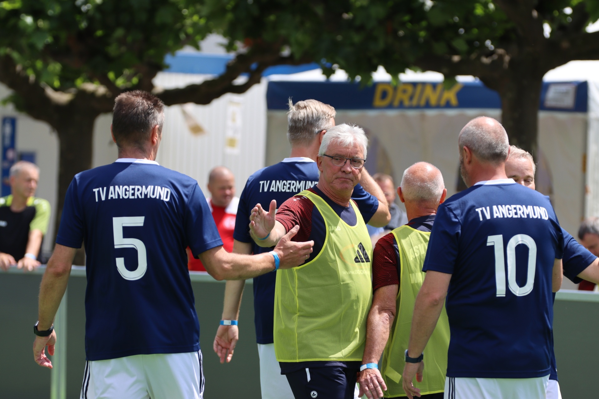 [{"clang_id":1,"value":"Drei Männer in Fußballtrikots \"TV Angermund\" (Nummern 5, 10 und ein Mann in gelber Weste) diskutieren auf einem Sportplatz. Im Hintergrund Zelte und Bäume."}]