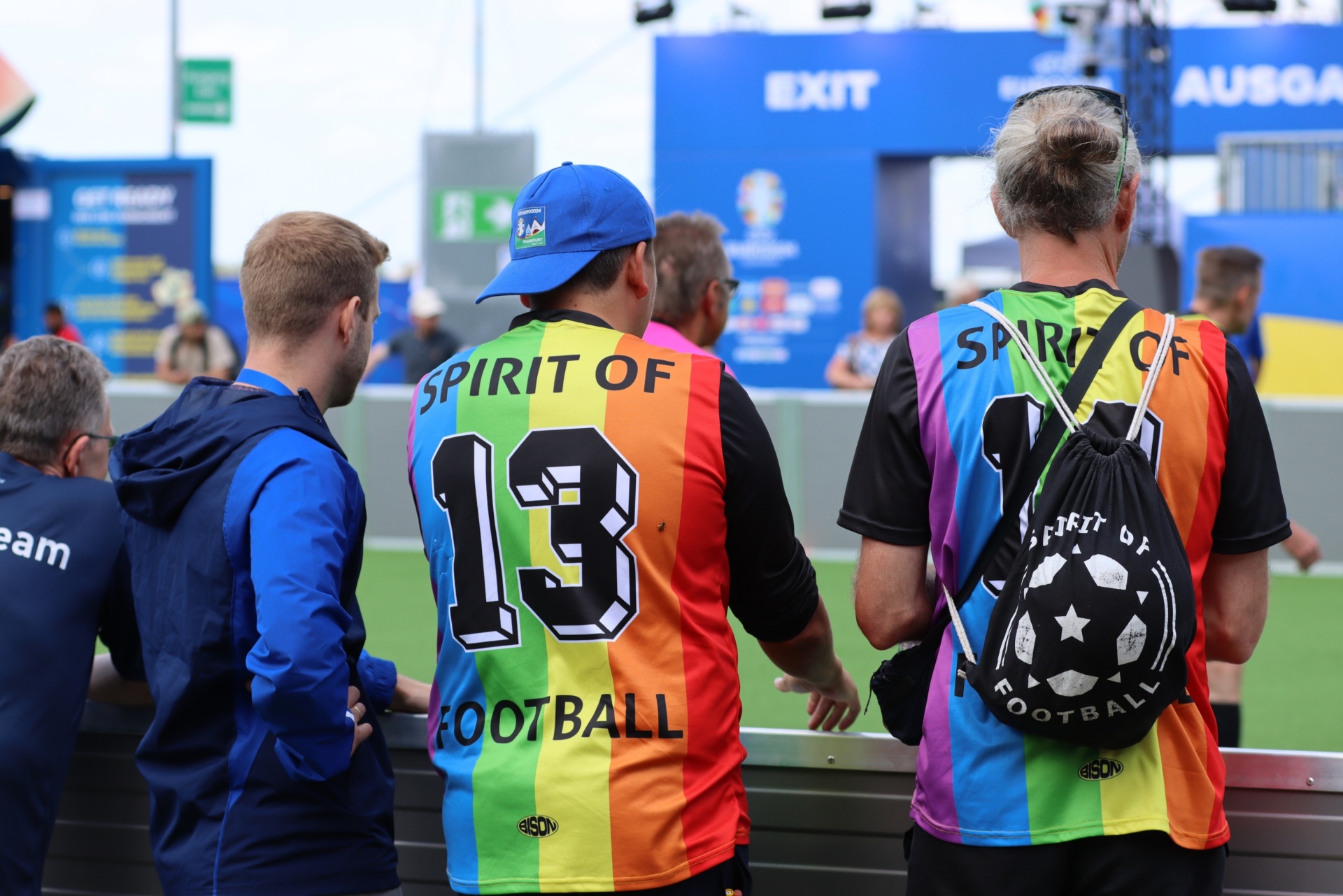 [{"clang_id":1,"value":"Drei Männer im Stadion. Zwei tragen bunte Trikots mit \"Spirit of Football\"-Aufschrift, einer blaues Teamkleid. Im Hintergrund ein Banner mit \"EXIT\"-Schriftzug."}]