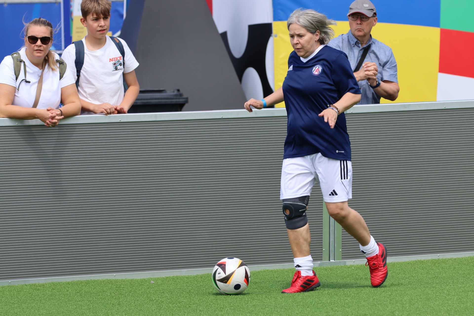 [{"clang_id":1,"value":"Frau mittleren Alters spielt Fußball auf grünem Rasen. Im Hintergrund stehen zwei Zuschauer und eine farbige Fahnenwand."}]