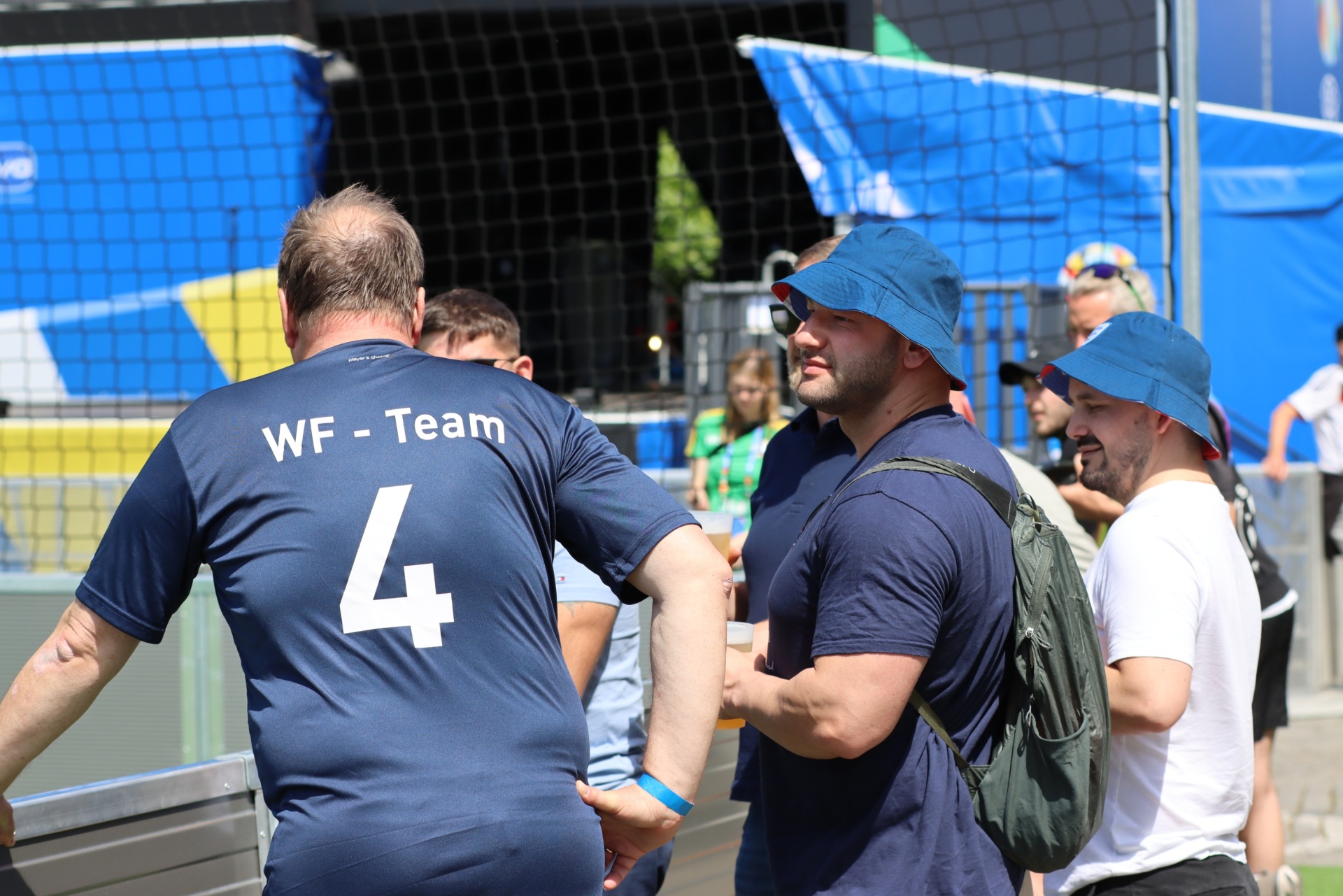 [{"clang_id":1,"value":"Drei Männer stehen draußen, vermutlich bei einem Sportevent. Ein Mann trägt ein blaues Trikot mit Aufschrift \"WF-Team\" und Nummer 4. Im Hintergrund ein Netz und Zuschauer."}]