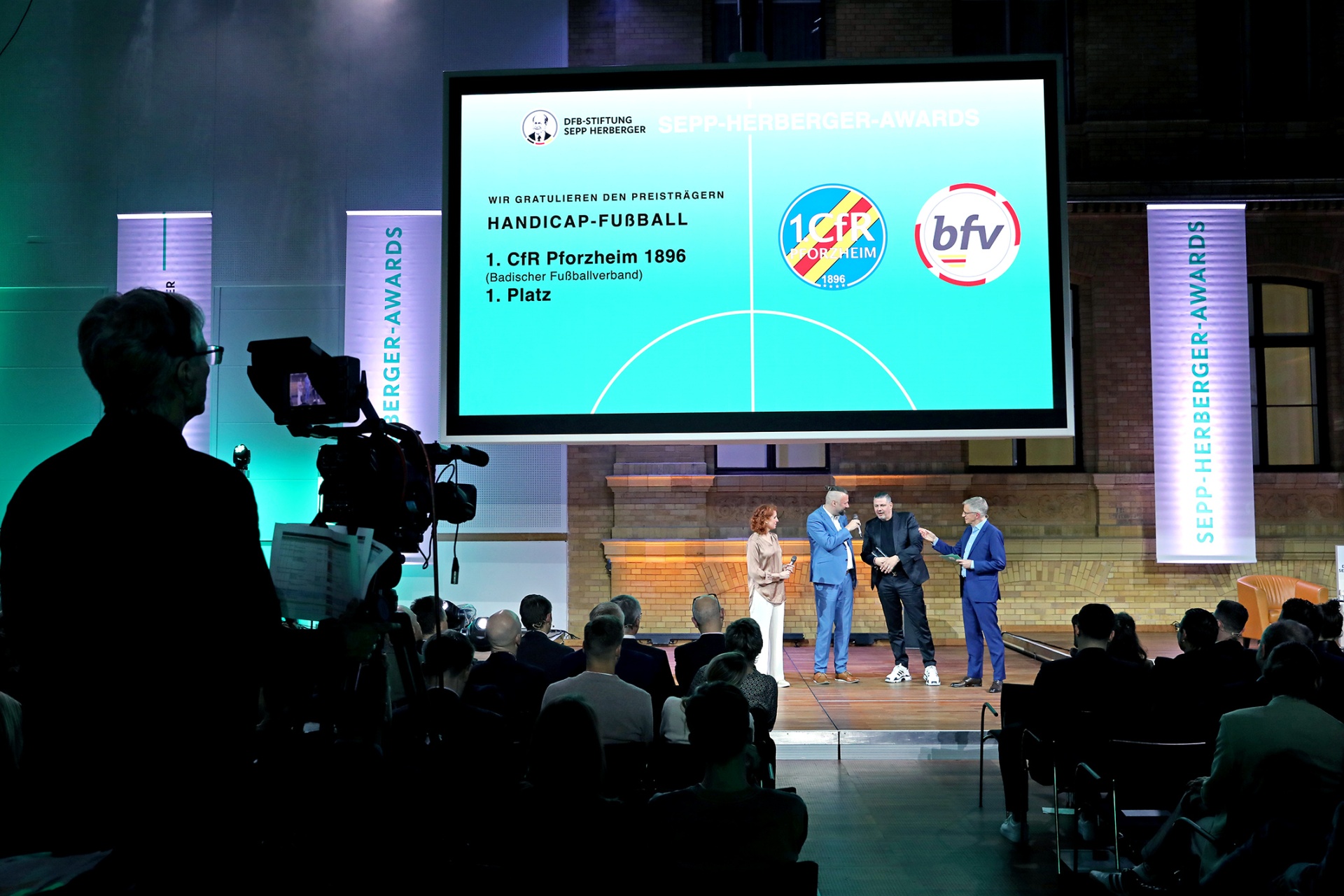 [{"clang_id":1,"value":"Preisverleihung: Bühne mit drei Personen, großem Bildschirm mit Text \"Handicap-Fußball, 1. CFR Pforzheim 1896, 1. Platz\". Publikum im Hintergrund."}]
