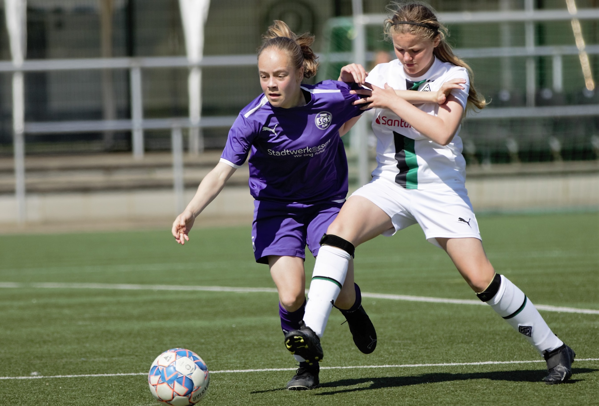 [{"clang_id":1,"value":"Zwei junge Fußballspielerinnen im Wettkampf. Links trägt eine Spielerin ein violettes Trikot, rechts ein weiß-grünes. Beide dribbeln den Ball auf einem grünen Rasenplatz."}]