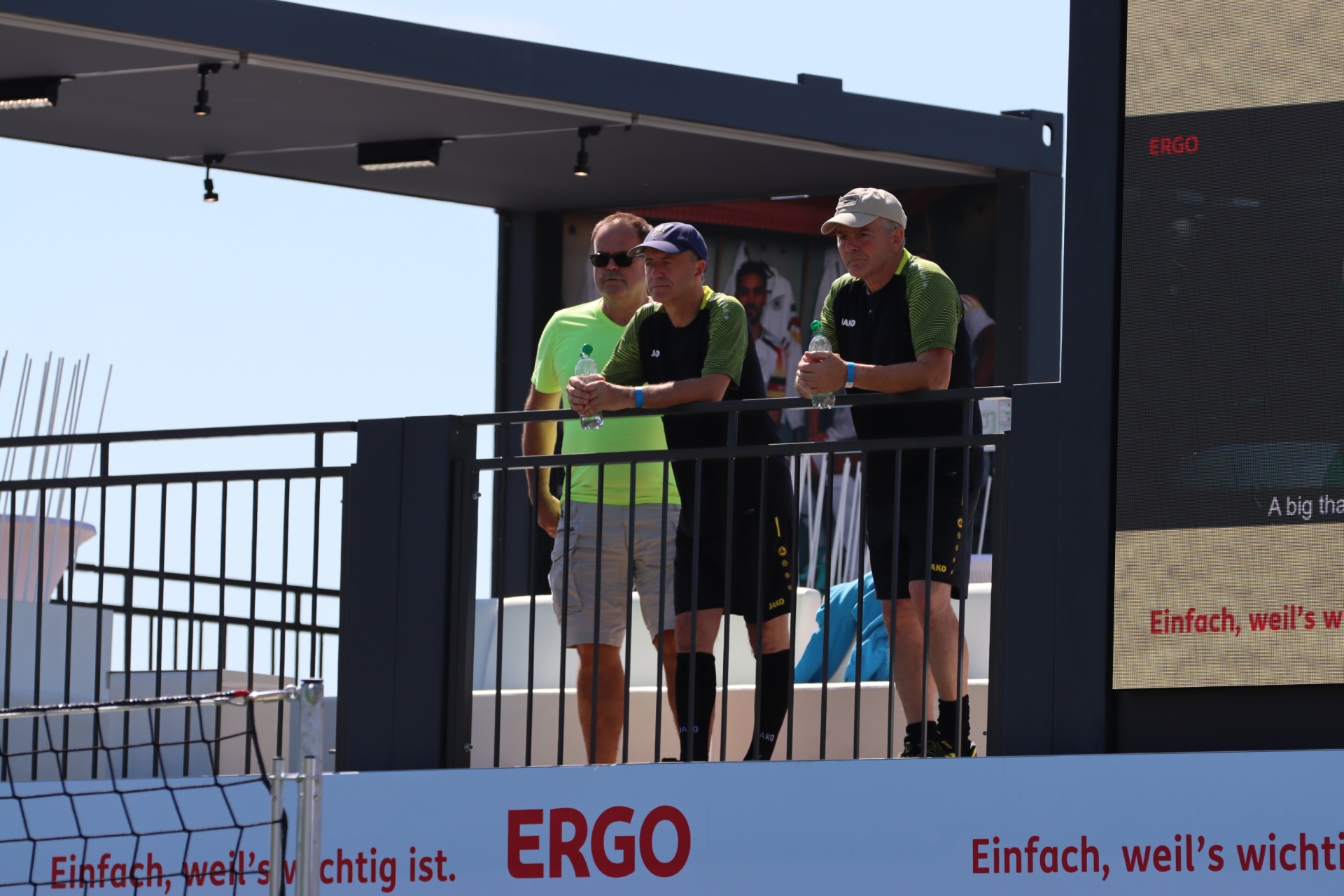 [{"clang_id":1,"value":"Drei Männer stehen an einer Absperrung vor einem Werbebanner mit dem Schriftzug \"ERGO\". Im Hintergrund eine große LED-Wand. Helles Tageslicht, blauer Himmel."}]