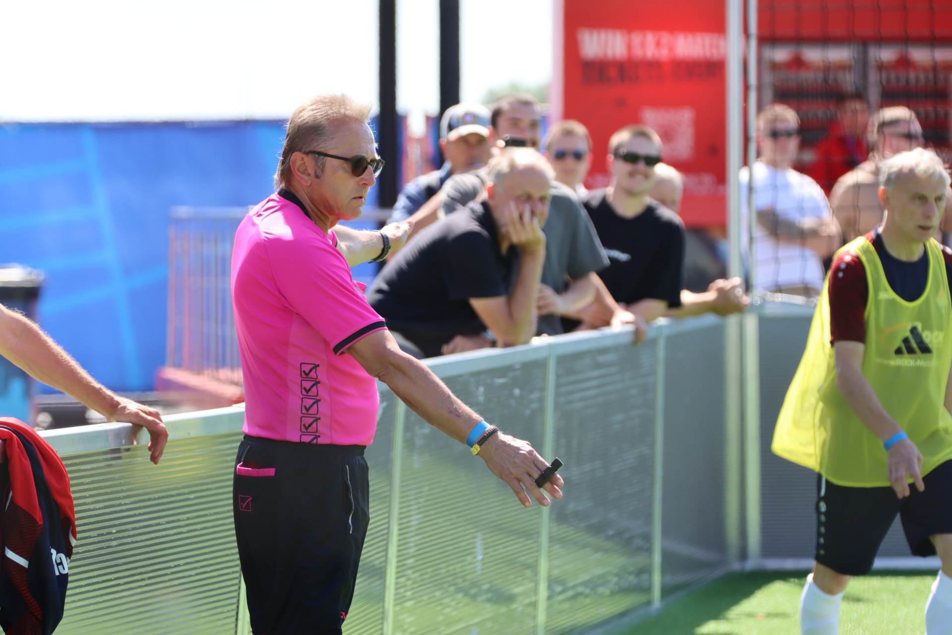 [{"clang_id":1,"value":"Fußballspiel: Schiedsrichter in Pink, mit Pfeife, am Spielfeldrand. Im Hintergrund Zuschauer und Spieler mit gelben Westen."}]