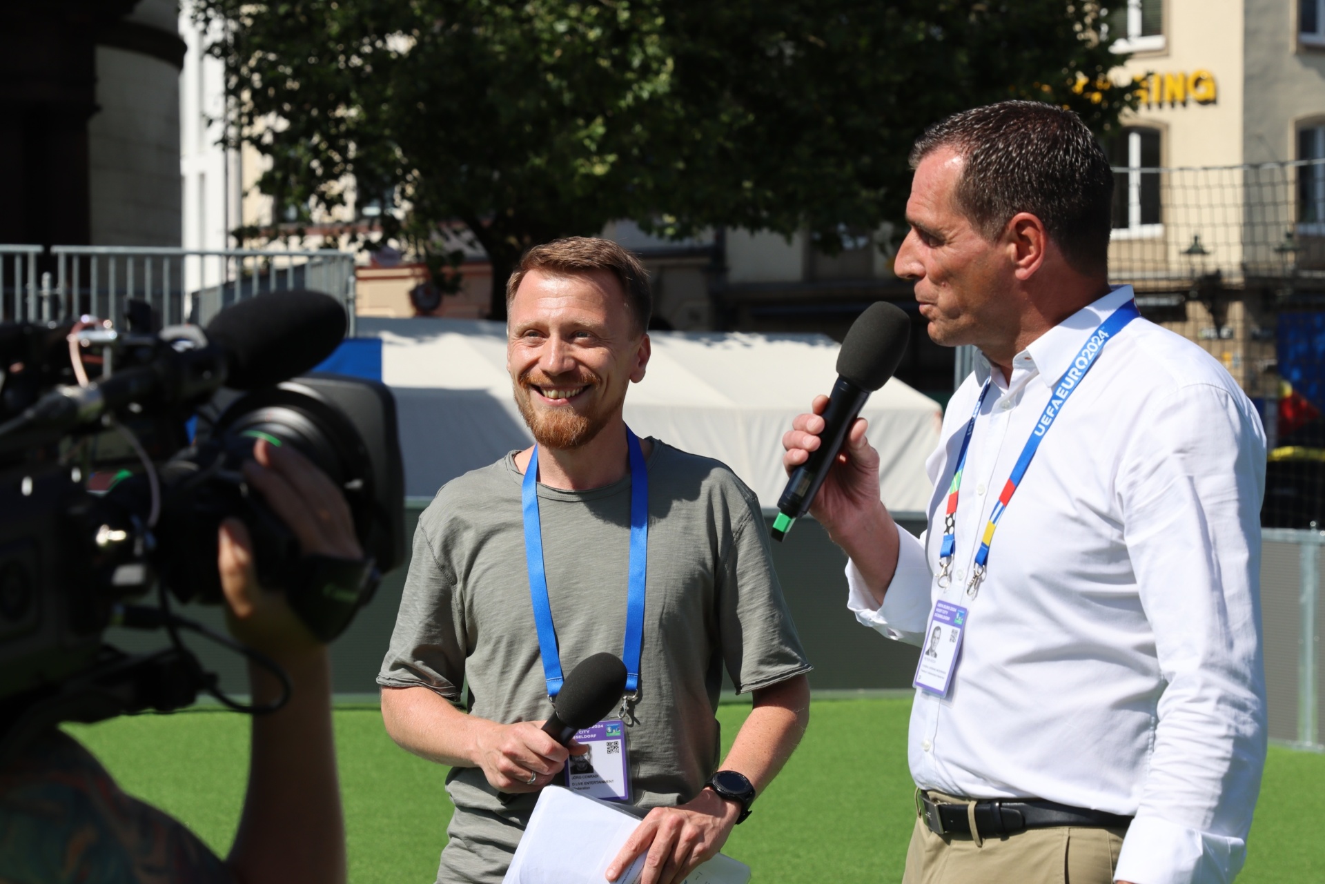 [{"clang_id":1,"value":"Zwei Männer werden interviewt. Links ein Mann mit Bart und grünem T-Shirt, rechts ein Mann mit Hemd und Mikrofon. Im Hintergrund Gebäude und eine Kamera."}]