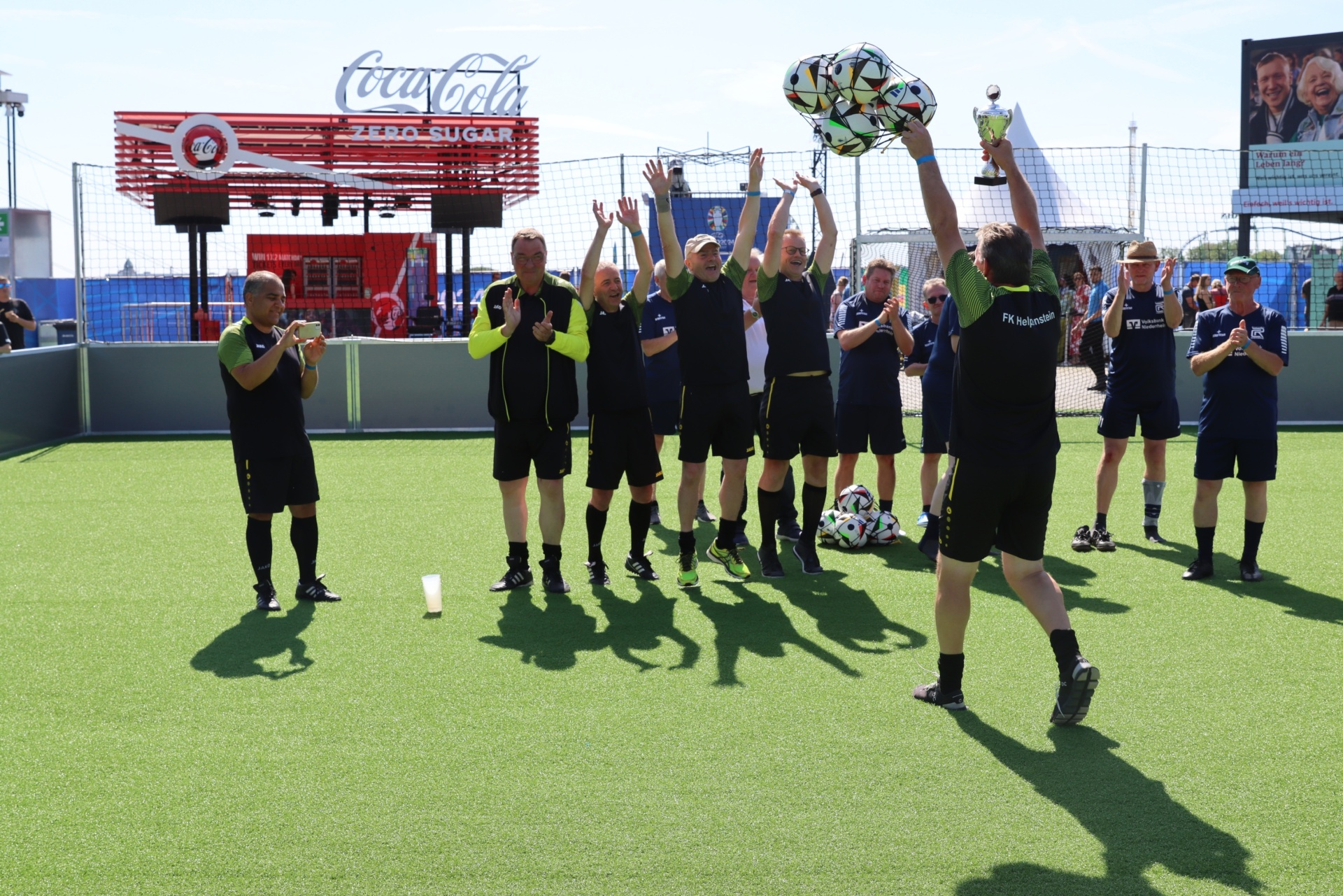 [{"clang_id":1,"value":"Fußballteam feiert Sieg. Ein Spieler hält den Pokal hoch, Teammitglieder applaudieren auf einem grünen Rasen vor einer Coca-Cola-Werbung."}]