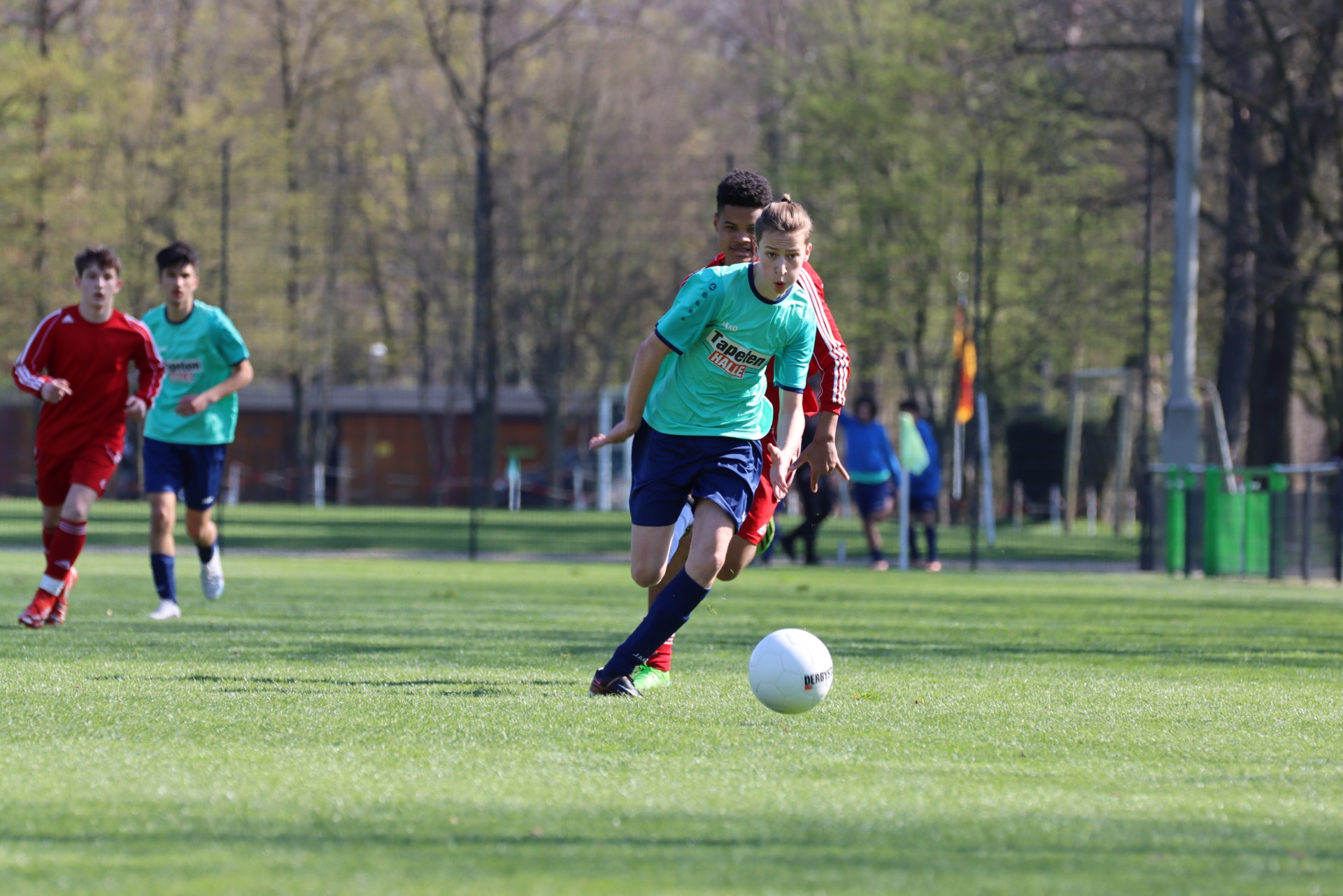 [{"clang_id":1,"value":"Jugendfußballspiel auf grünem Rasenfeld. Spieler in roten und türkisfarbenen Trikots, ein Spieler dribbelt den Ball. Bäume im Hintergrund."}]