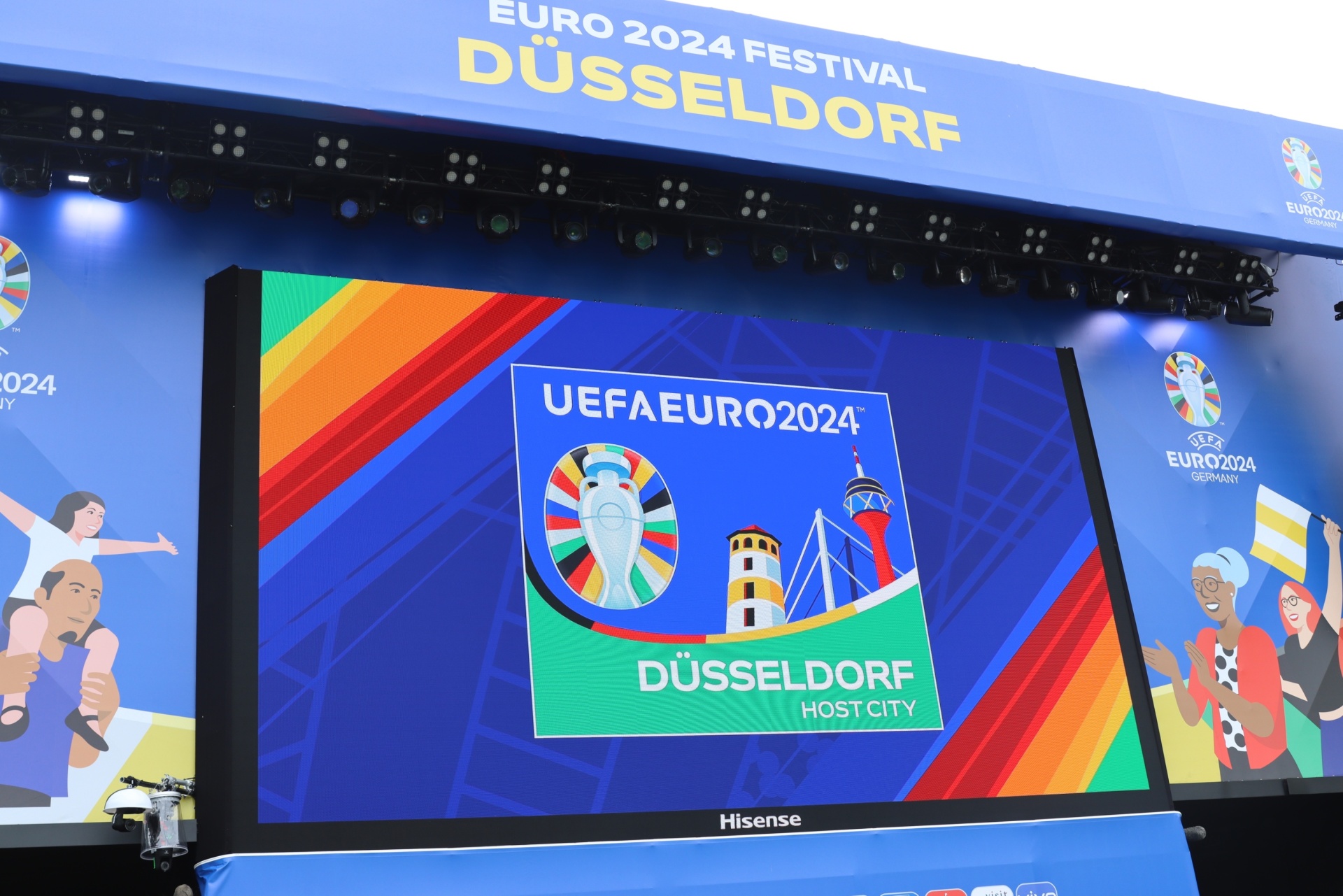 [{"clang_id":1,"value":"EURO 2024 Festival Düsseldorf: Großes LED-Display zeigt Turnierlogo mit Skyline, darunter \"HOST CITY\". Bunte Banner mit stilisierten Personen im Hintergrund."}]