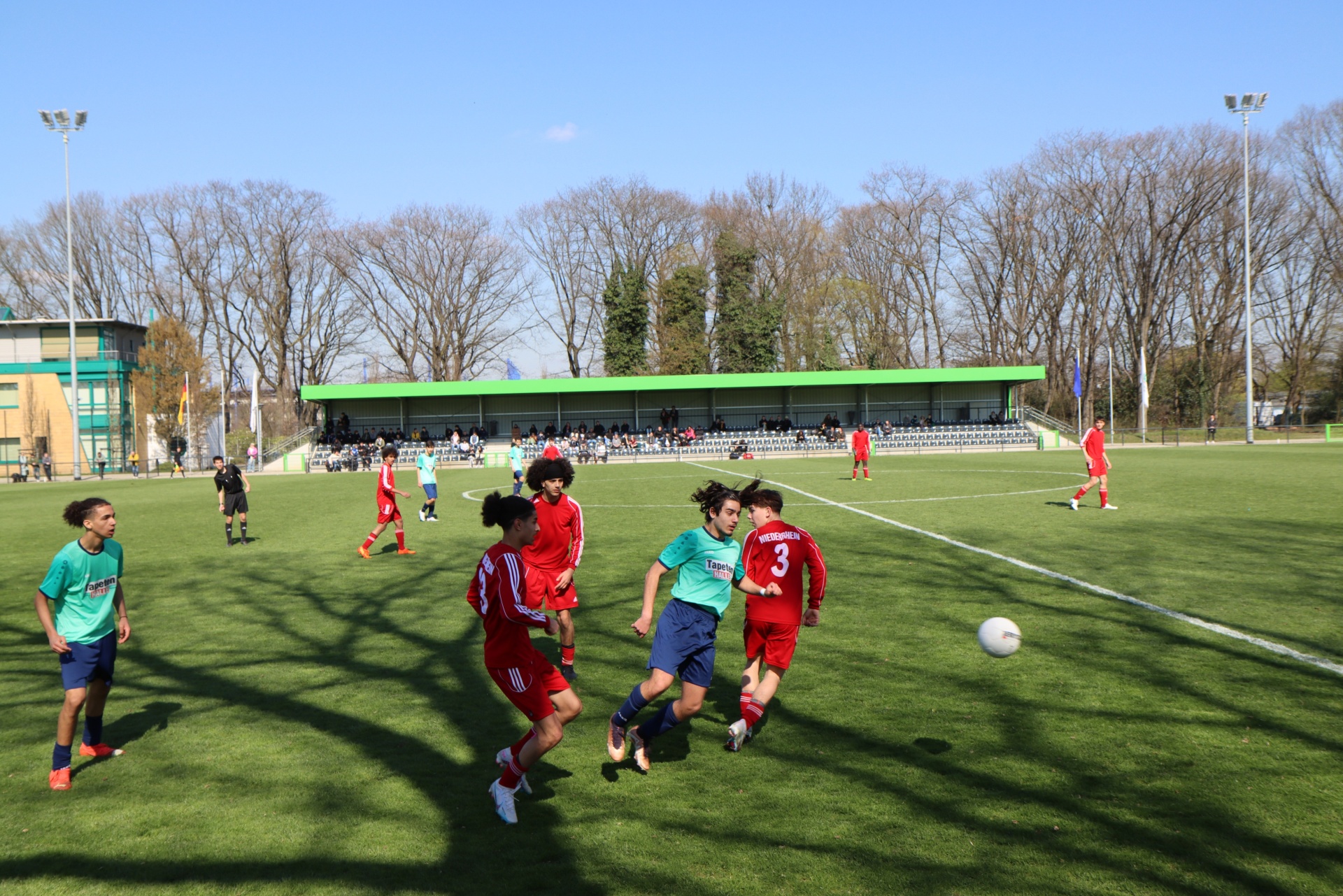 [{"clang_id":1,"value":"Fußballspiel auf Rasenplatz. Spieler in rot und blau/grün kämpfen um den Ball. Im Hintergrund Zuschauer und Bäume unter blauem Himmel."}]