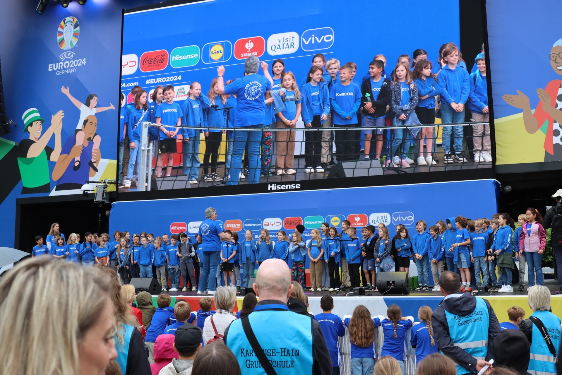 [{"clang_id":1,"value":"Chor von Kindern in blauen T-Shirts steht auf einer Bühne vor einer großen Leinwand mit EURO2024-Logo und Sponsoren. Publikum im Hintergrund."}]