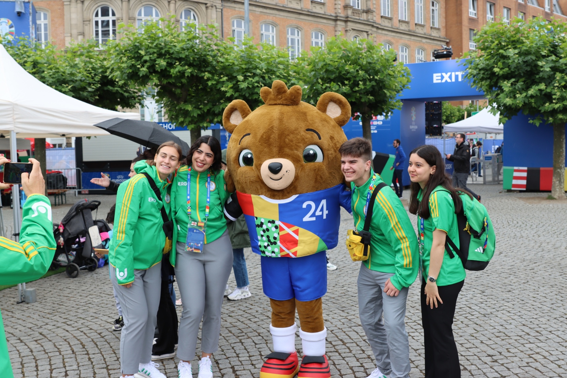 [{"clang_id":1,"value":"Personengruppe mit Maskottchen \"Malik\" (brauner Bär mit Fußballtrikot Nr. 24) vor Gebäudefassade. Einige tragen grüne Jacken mit Logo. Im Hintergrund Zelte und Menschen."}]