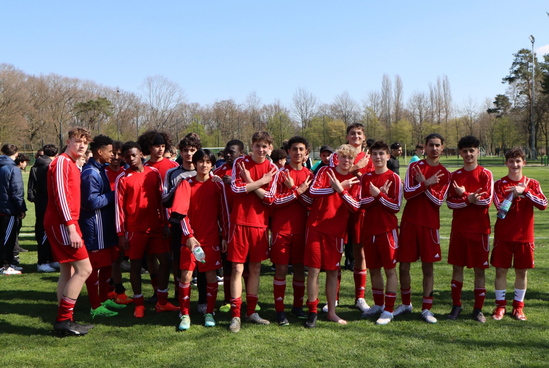 [{"clang_id":1,"value":"Fußballmannschaft in roten Trikots und weißen Hosen steht auf grünem Rasen. Einige Spieler haben die Arme verschränkt. Im Hintergrund Bäume und andere Spieler."}]