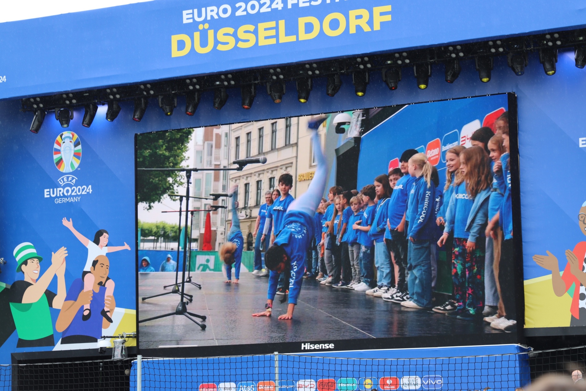 [{"clang_id":1,"value":"Große LED-Wand mit Video von Kindern in blauen Sportanzügen, die eine Turnvorführung in einer Stadt zeigen. Hintergrund: \"EURO 2024 FESTIVAL DÜSSELDORF\"."}]