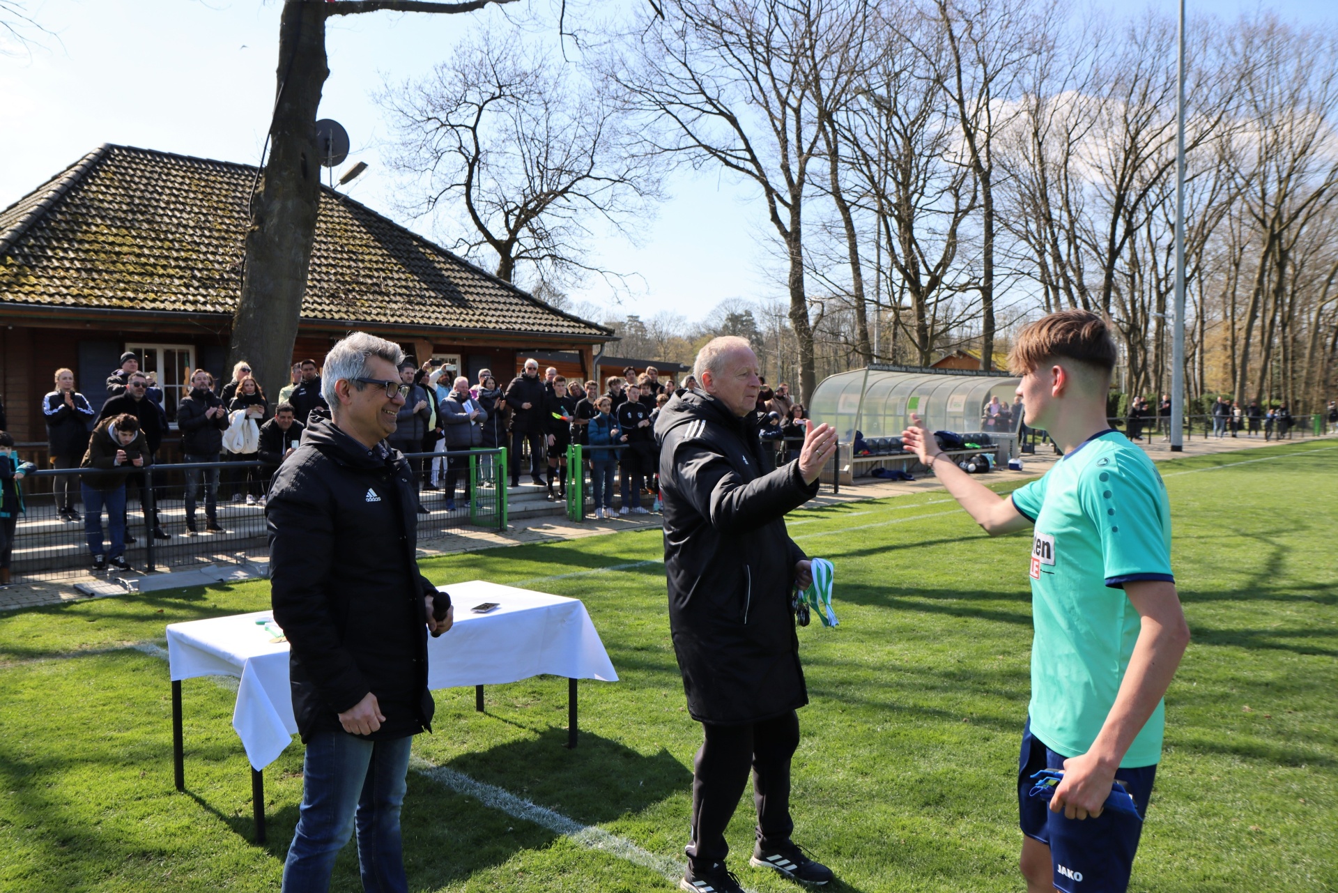[{"clang_id":1,"value":"Drei Männer stehen auf einem Fußballfeld vor einem Tisch mit weißer Tischdecke. Im Hintergrund ein Clubhaus und Zuschauer."}]