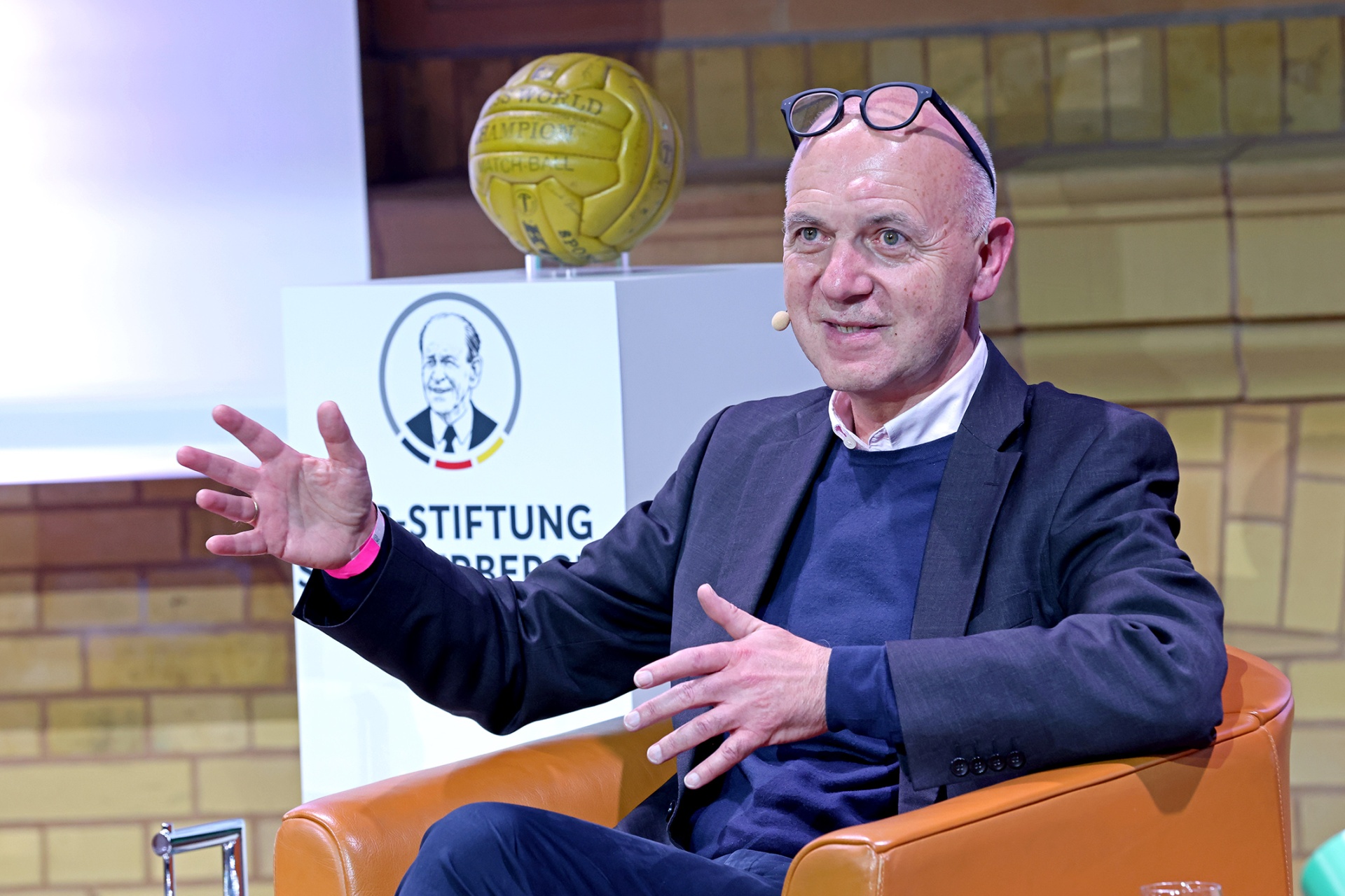 [{"clang_id":1,"value":"Mann mit Brille sitzt in einem Sessel, gestikuliert und spricht. Im Hintergrund eine Ziegelwand und ein Sockel mit Fußball und Logo \"Stiftung\"."}]