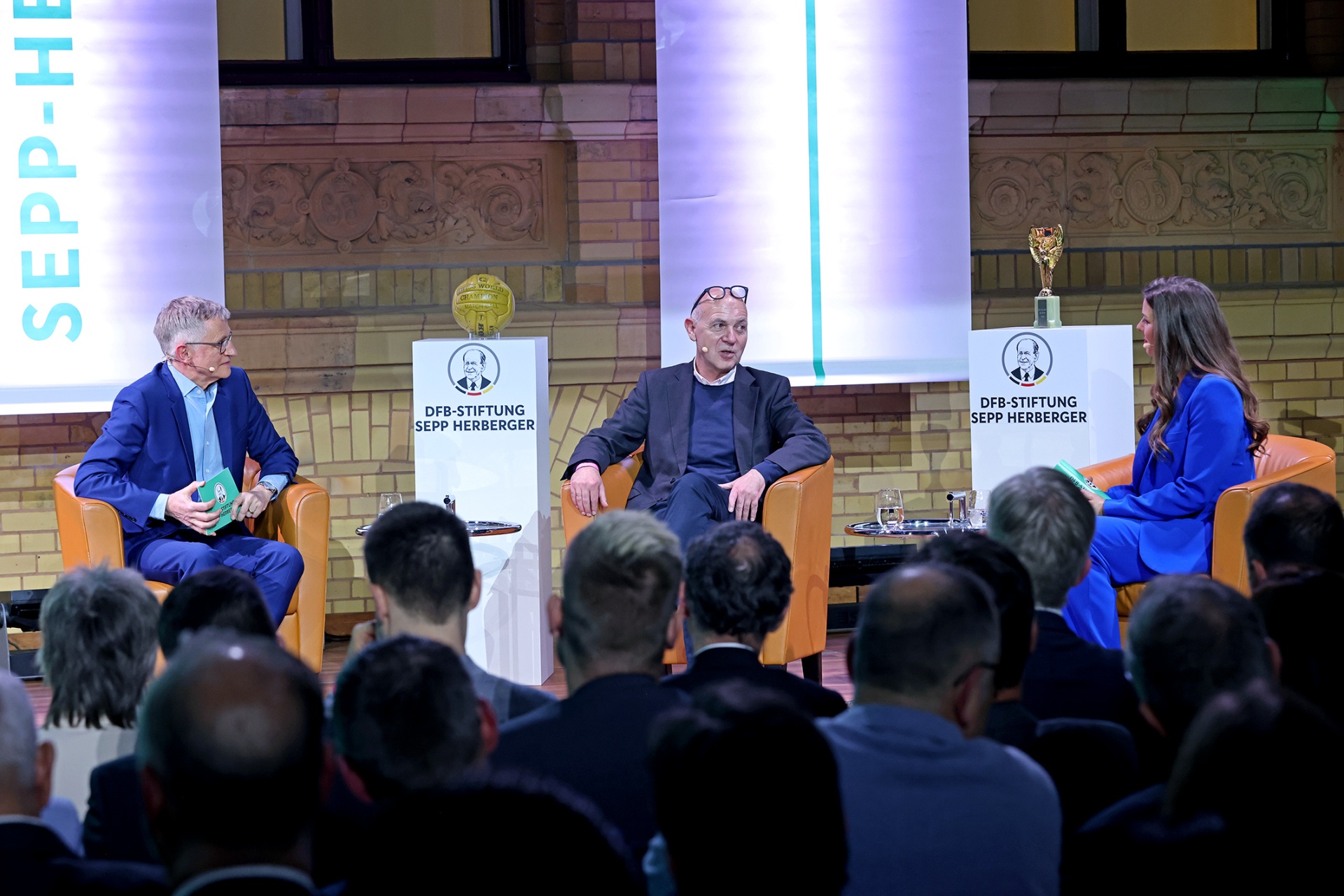 [{"clang_id":1,"value":"Panelsitzung mit drei Personen auf Bühne vor Publikum. Links und rechts Logos der \"DFB-Stiftung Sepp Herberger\". Im Hintergrund dekorative Wandgestaltung."}]