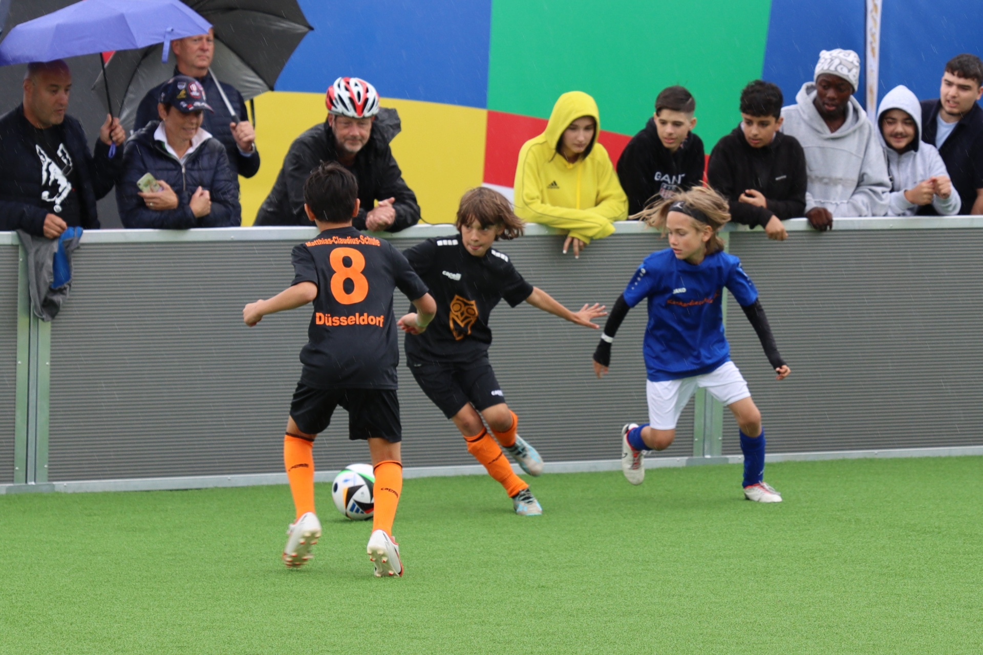 [{"clang_id":1,"value":"Drei Kinder spielen Fußball auf einem grünen Kunstrasenplatz. Im Hintergrund Zuschauer unter Regenschirmen und mit Regenbekleidung."}]