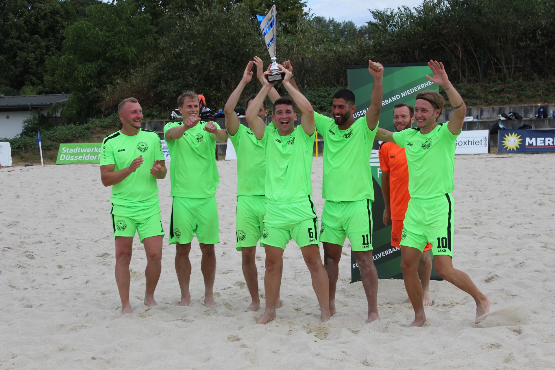 [{"clang_id":1,"value":"Sieben Fussballspieler in leuchtend grünen Trikots feiern auf Sandstrand Sieg mit Pokal. Im Hintergrund Werbebanner und Publikum."}]