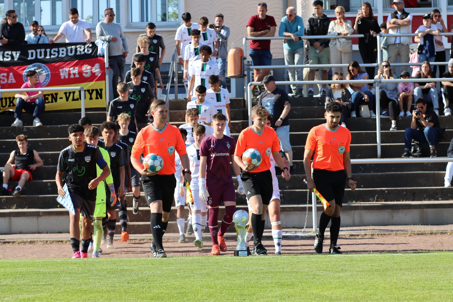 [{"clang_id":1,"value":"Fußballspieler in verschiedenen Trikots laufen auf das Spielfeld. Im Hintergrund Zuschauer auf einer Tribüne und eine Stadionbande mit Aufschrift."}]