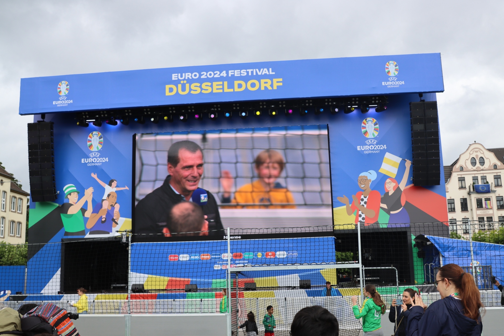 [{"clang_id":1,"value":"EURO 2024 Festival Düsseldorf: Großbildschirm zeigt Fußballübertragung mit zwei Männern und zwei Kindern. Publikum steht davor. Im Hintergrund Gebäude."}]