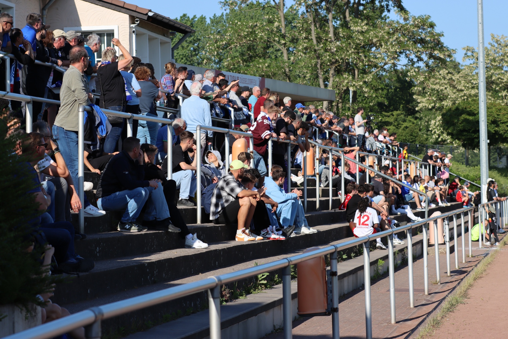[{"clang_id":1,"value":"Zuschauer auf einer Stadiontribüne bei einem Fußballspiel. Viele Menschen sitzen und stehen dicht gedrängt, überwiegend in Freizeitkleidung. Im Hintergrund Bäume und ein Gebäude."}]
