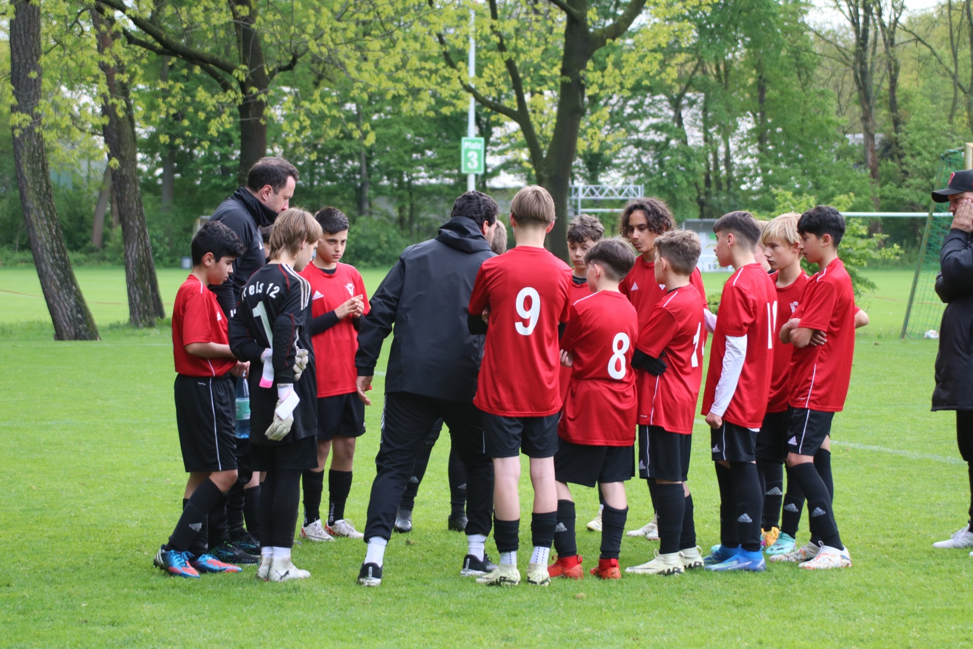 [{"clang_id":1,"value":"Fußballteam im Kreis um Trainer auf grünem Rasenfeld. Spieler tragen rote und schwarze Trikots. Bäume im Hintergrund."}]