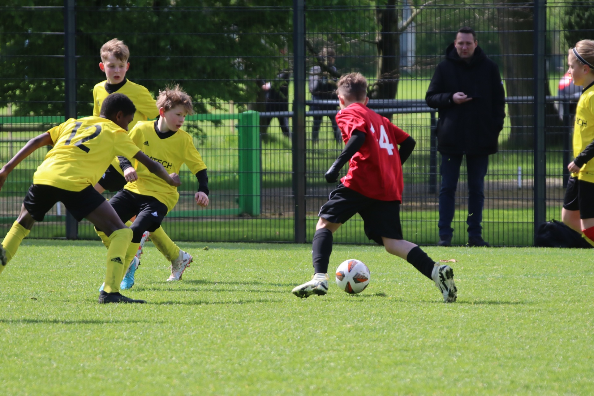 [{"clang_id":1,"value":"Jugendfußballspiel: Zwei Spieler in gelben Trikots verfolgen einen Spieler in rotem Trikot mit Ball auf grünem Rasen. Im Hintergrund steht ein Zuschauer."}]