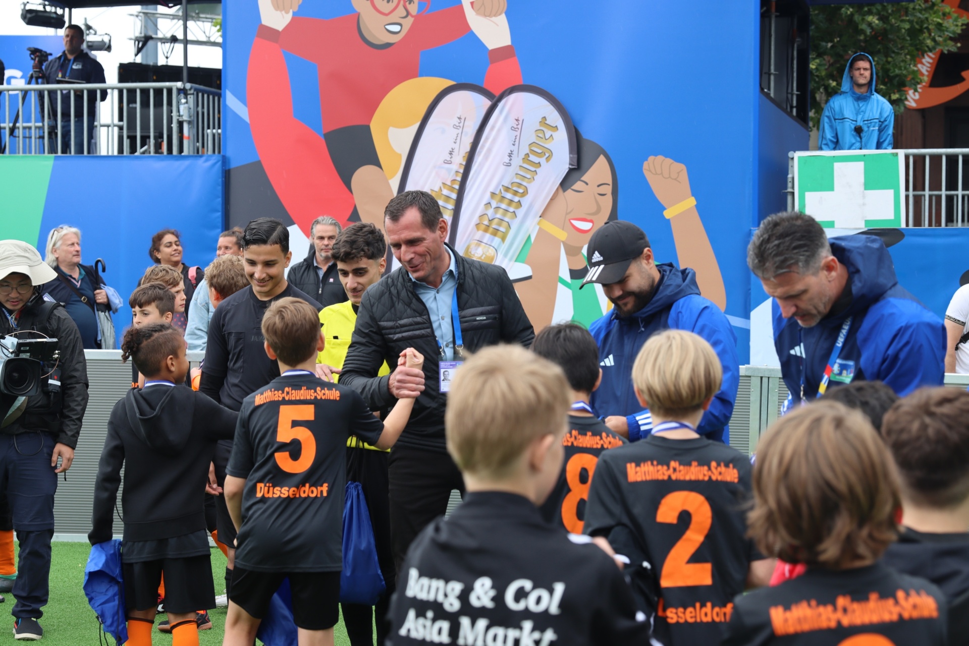 [{"clang_id":1,"value":"Fußballspieler-Kinder in schwarzen Trikots mit Schriftzug \"Matthias-Claudius-Schule Düsseldorf\" werden von Erwachsenen begrüßt. Im Hintergrund eine bunte Wand und Zuschauertribüne."}]
