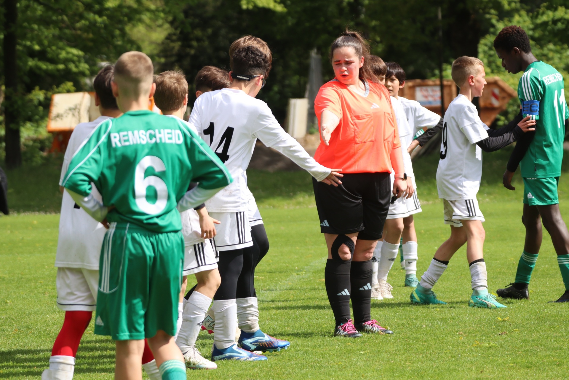 [{"clang_id":1,"value":"Fußballspiel: Gruppe Jugendlicher in Sportkleidung, einige mit Trikots (Nummern 6, 14), und eine Schiedsrichterin diskutieren auf dem Fußballfeld."}]
