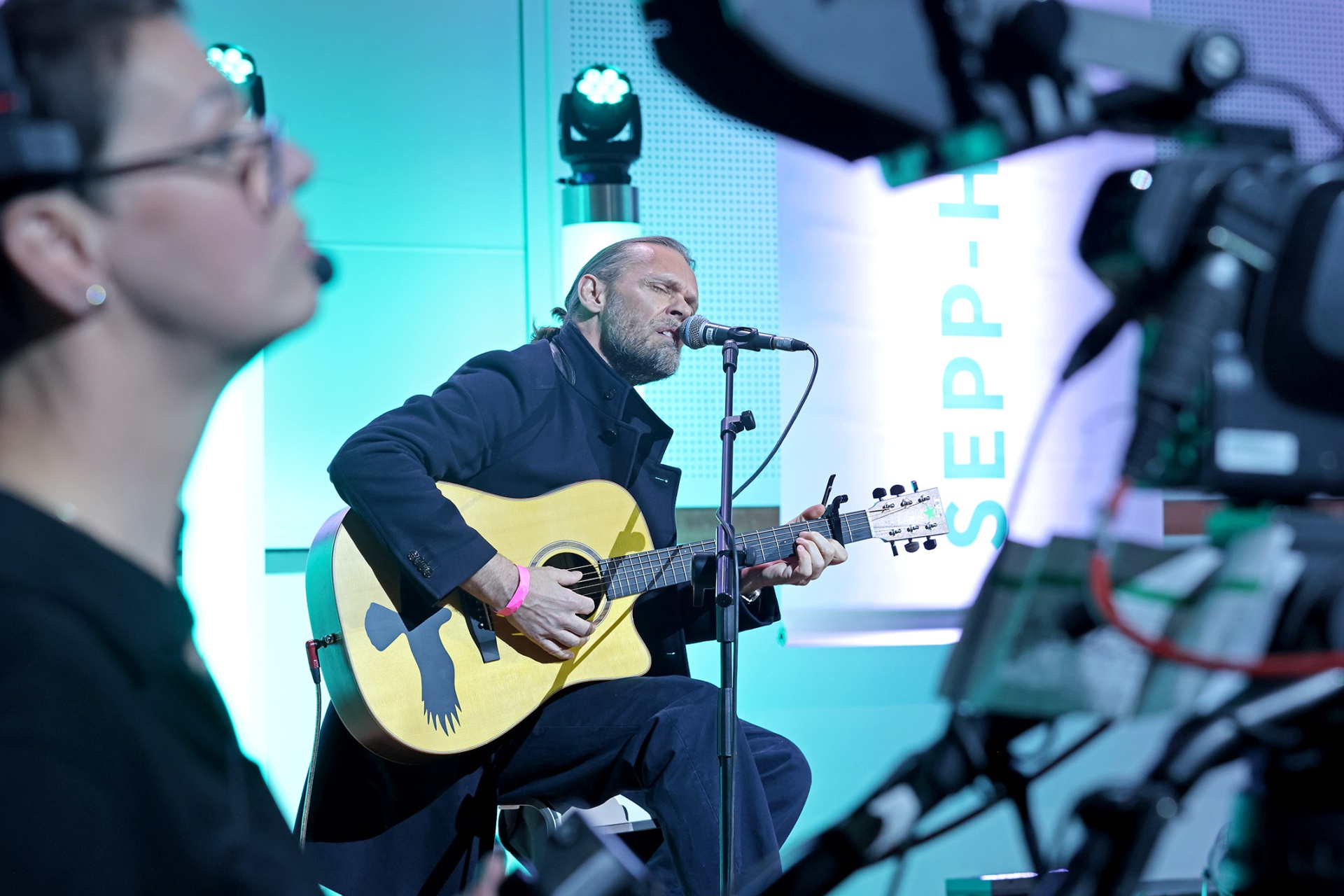 [{"clang_id":1,"value":"Mann mit Gitarre singt in ein Mikrofon. Im Hintergrund eine Person mit Headset und Kameras. Lichteffekte in Blau und Weiß."}]
