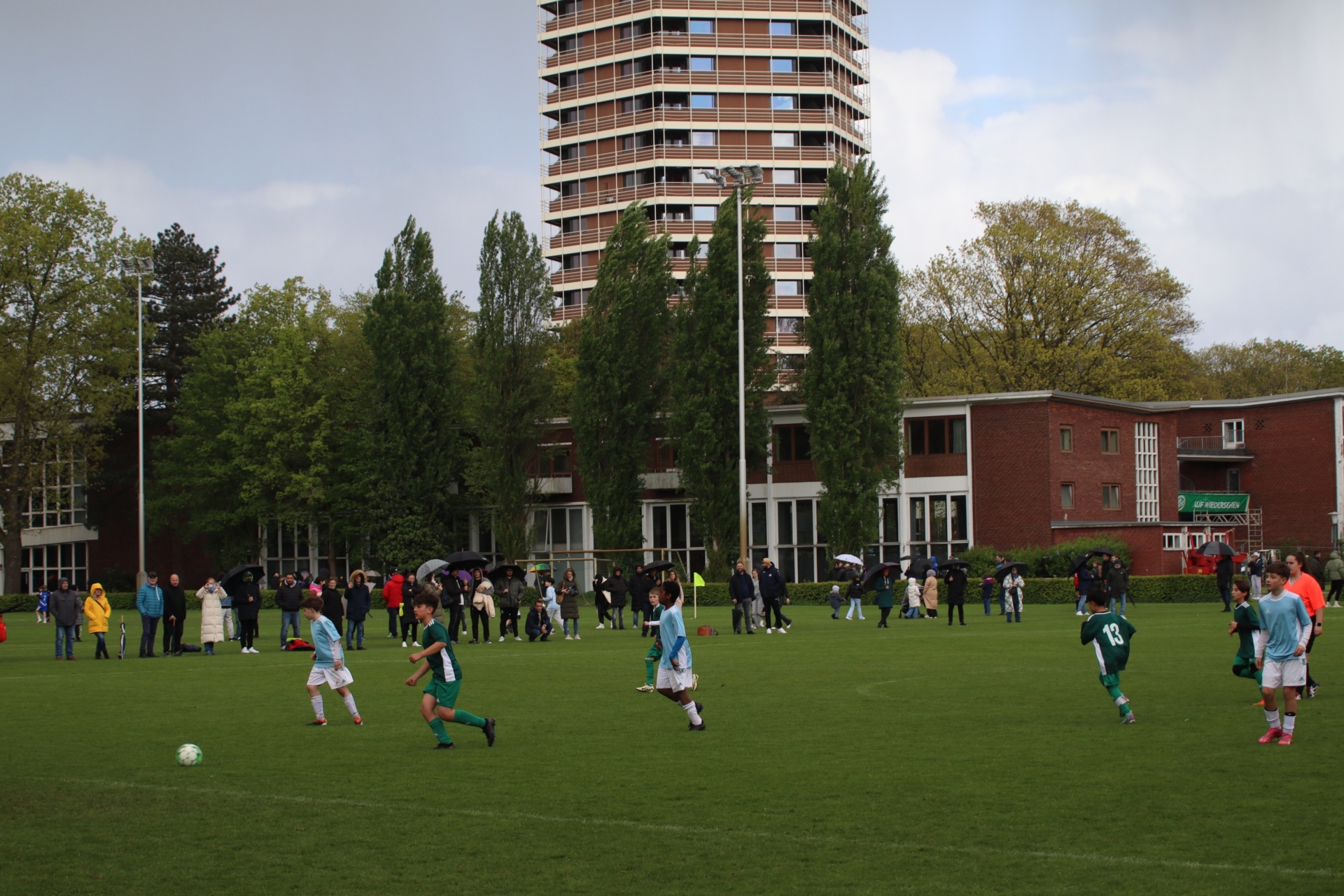 [{"clang_id":1,"value":"Fußballspiel auf grünem Rasen. Im Hintergrund ein hoher Wohnblock und Bäume. Zuschauer stehen am Spielfeldrand."}]