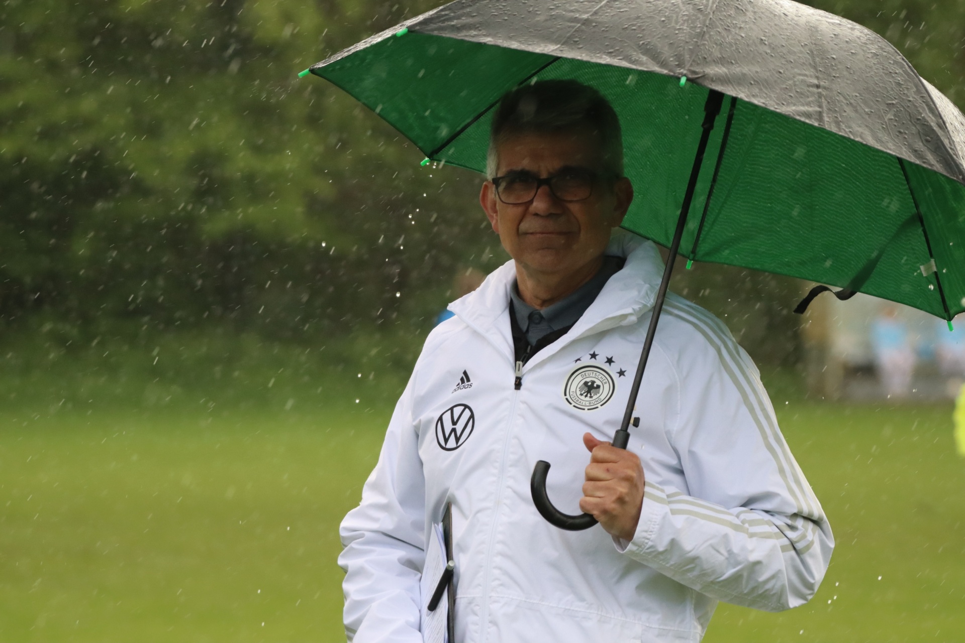 [{"clang_id":1,"value":"Mann mit Brille und weißer Sportjacke hält grünen Regenschirm im Regen auf einem Fußballfeld. Im Hintergrund verschwommen andere Personen und Grünflächen."}]