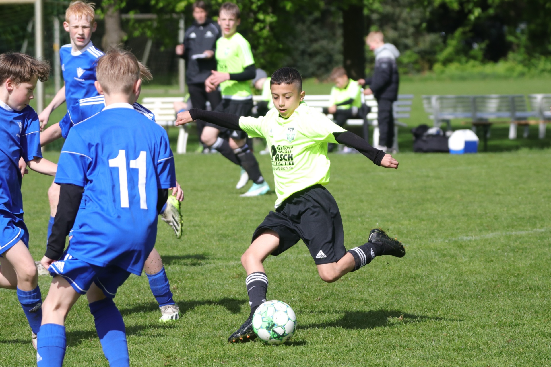 [{"clang_id":1,"value":"Fußballspiel: Junge in gelbem Trikot schießt auf das Tor. Mehrere Spieler in blauen Trikots um ihn herum auf einem Rasenplatz. Bäume im Hintergrund."}]
