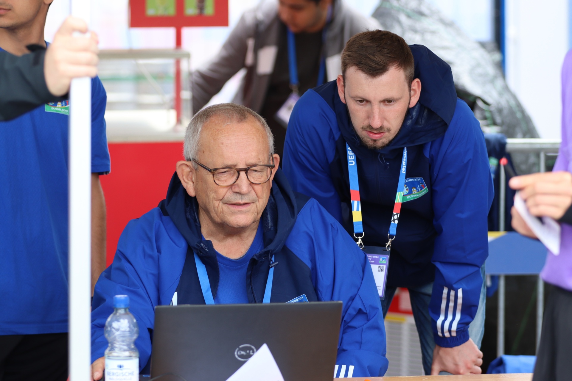 [{"clang_id":1,"value":"Zwei Männer an einem Tisch. Älterer Mann mit Brille tippt auf Laptop, jüngerer Mann schaut über seine Schulter. Beide tragen blaue Trainingsjacken mit UEFA-Ausweisen."}]