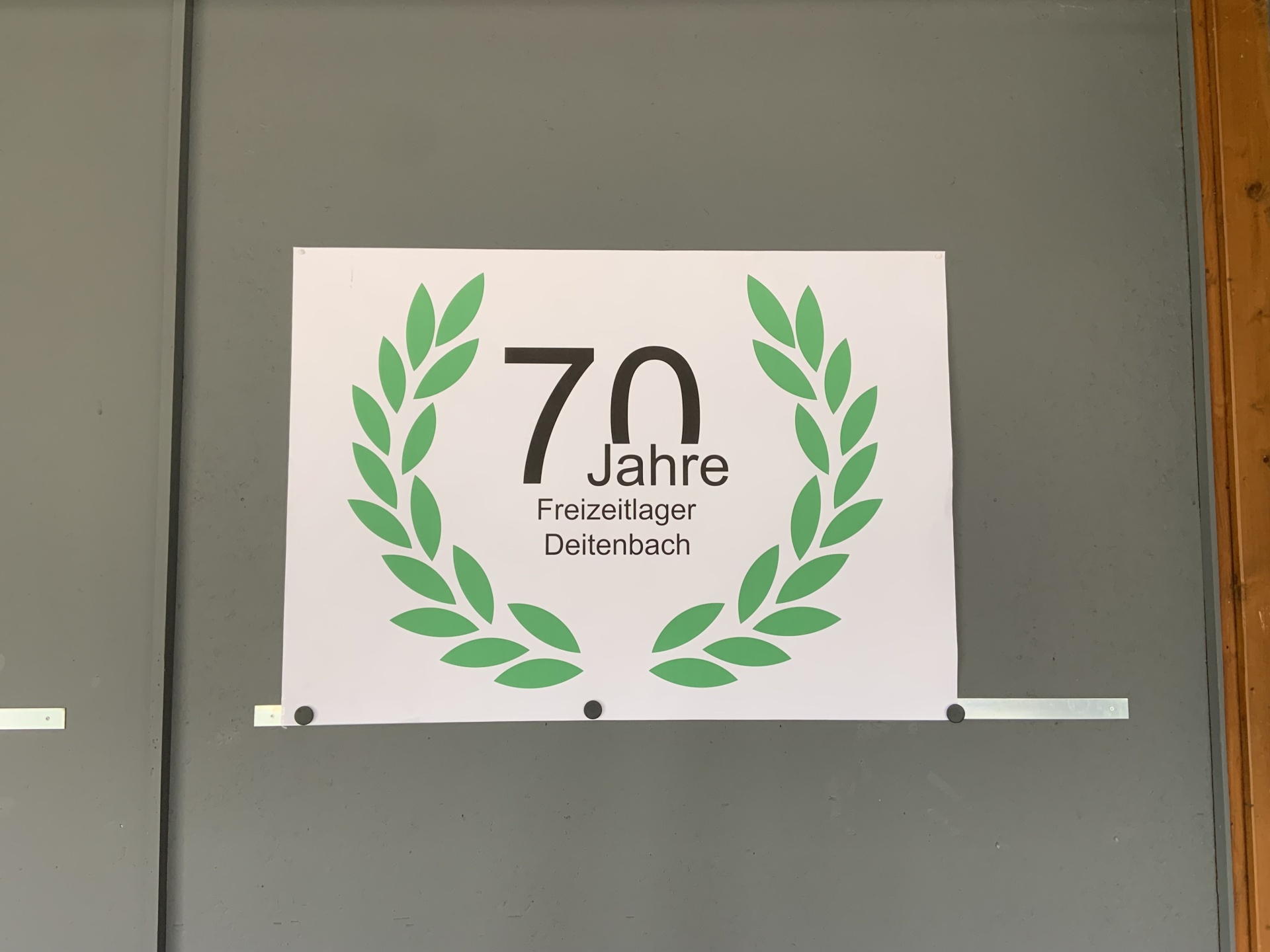 [{"clang_id":1,"value":"Weißes Plakat mit grüner Schrift: \"70 Jahre\" und darunter \"Freizeitlager Deitenbach\", umgeben von Lorbeerkränzen. Angebracht an grauer Wand."}]