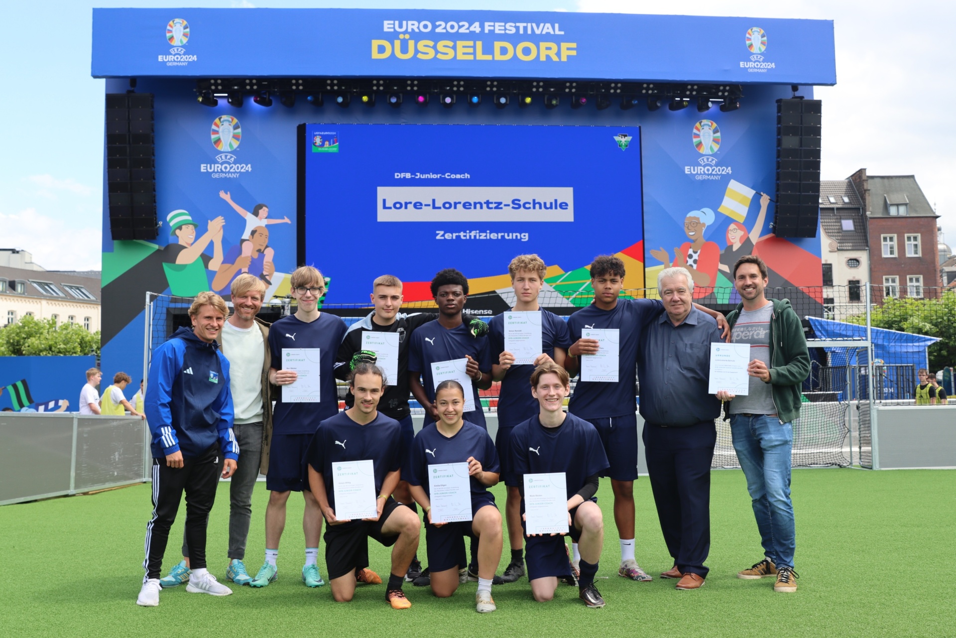 [{"clang_id":1,"value":"Gruppe Jugendlicher mit Zertifikaten vor LED-Wand mit EURO 2024 Logo und Text \"Lore-Lorentz-Schule Zertifizierung\". Erwachsene stehen am Rand. Fußballfeld im Hintergrund."}]