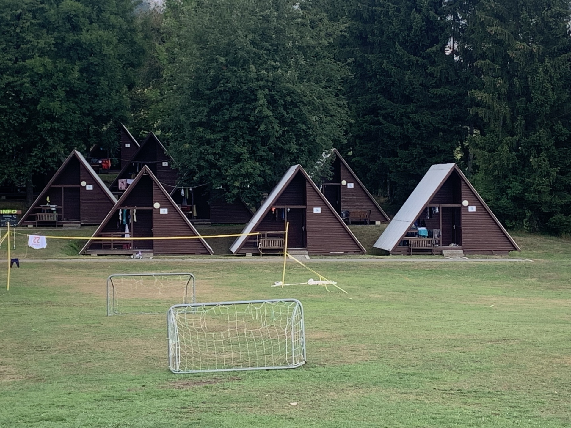 [{"clang_id":1,"value":"Dunkler Hintergrund mit fünf A-förmigen Holzhütten auf einer Wiese. Vor den Hütten ein Fußballtornetz. Im Hintergrund dichter Wald."}]