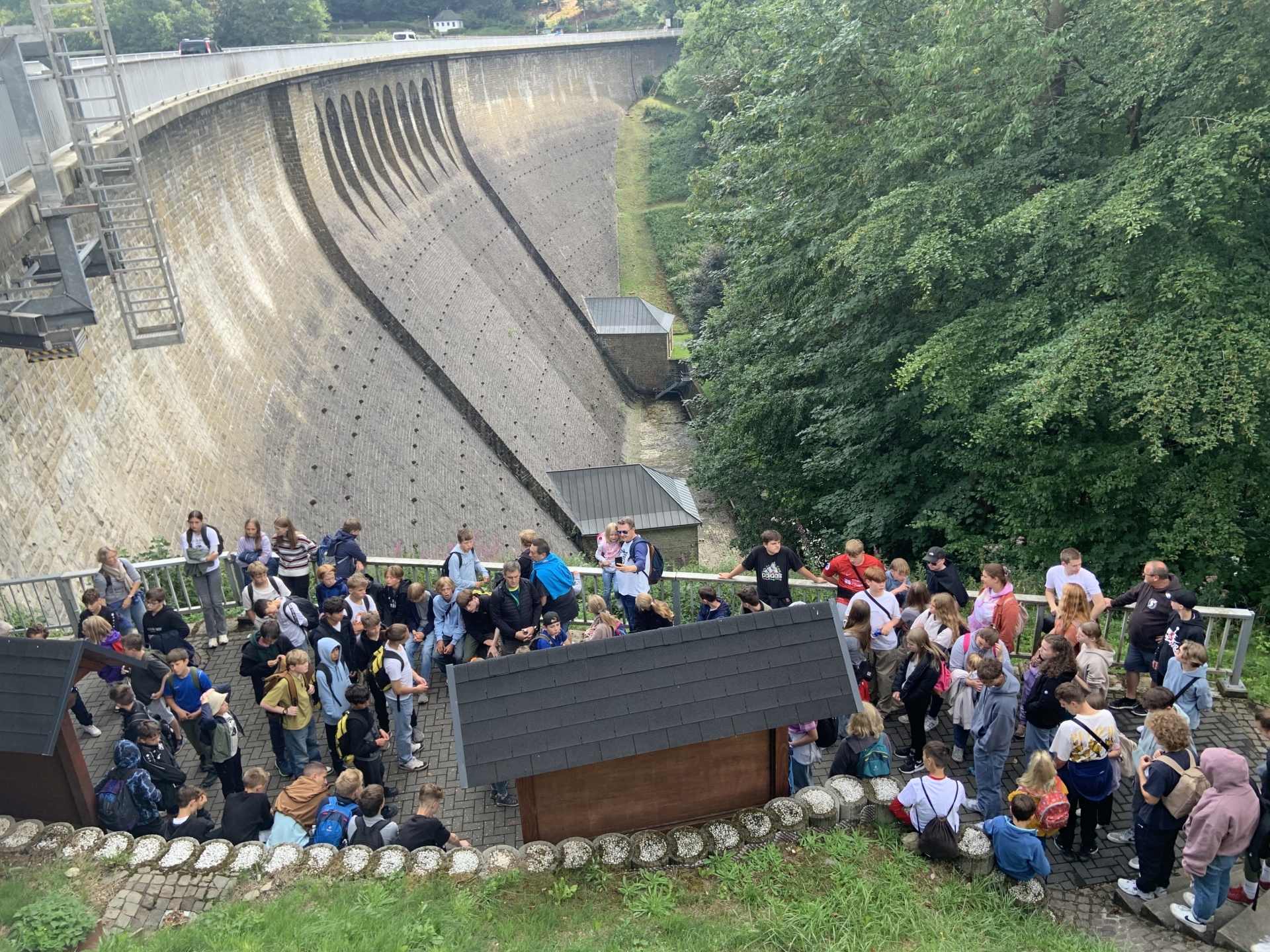 [{"clang_id":1,"value":"Große Gruppe Menschen vor einer hohen, gebogenen Stauanlage aus Beton. Rechts dichter Wald, im Hintergrund Häuser."}]